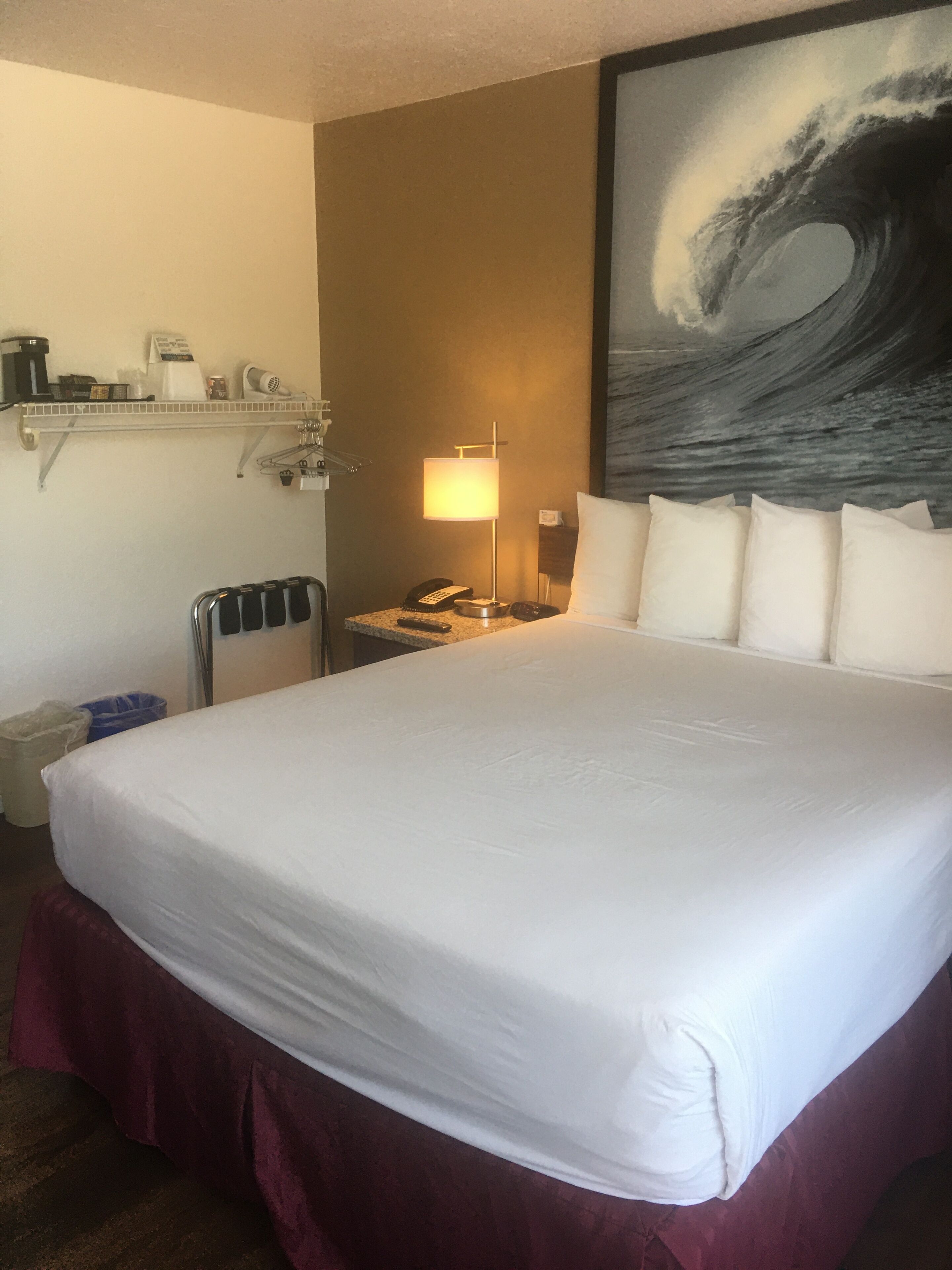 A hotel room with a large bed, a nightstand with a lamp, a phone, and a painting of a wave on the wall.