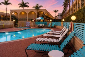 Une piscine avec des chaises longues et des parasols, un chemin pavé et un bâtiment doté d’arches et de balcons.
