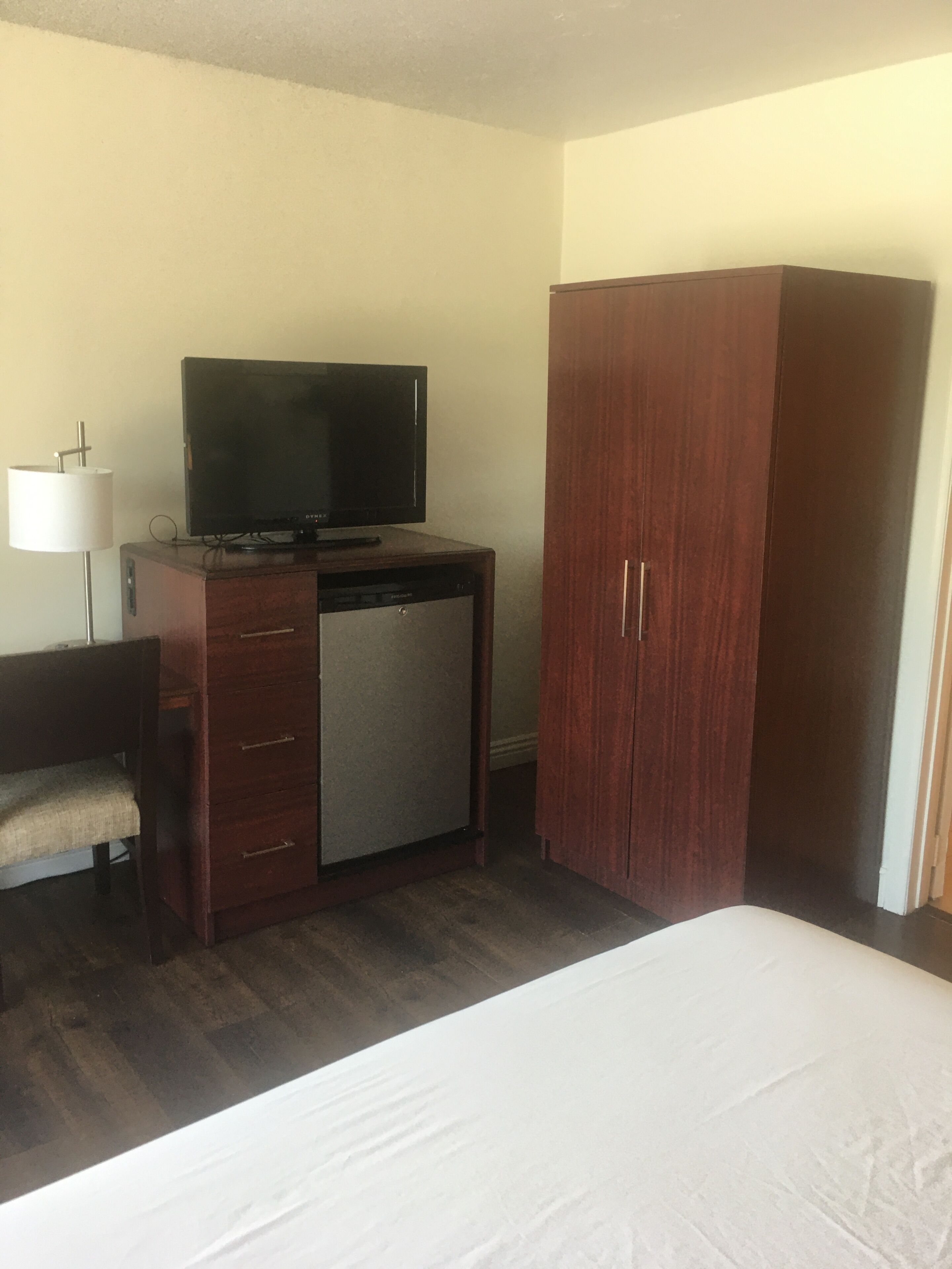 A hotel room with a bed, a desk with a television, a wardrobe, and a lamp.