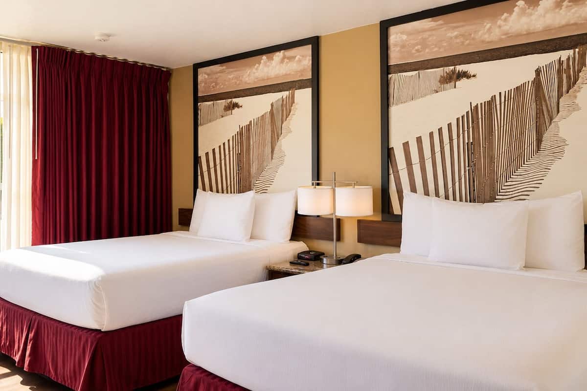 A hotel room with two beds, a window with a view of greenery, and artwork on the walls.