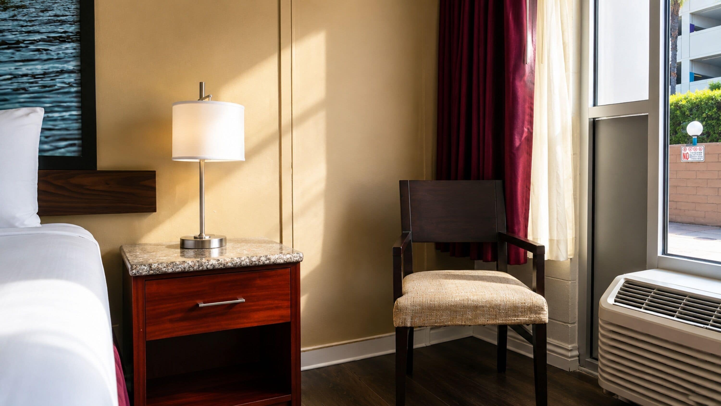 Una habitación de hotel con una cama, una mesita de noche con lámpara, una silla, una ventana con cortinas y un aire acondicionado instalado en la pared.