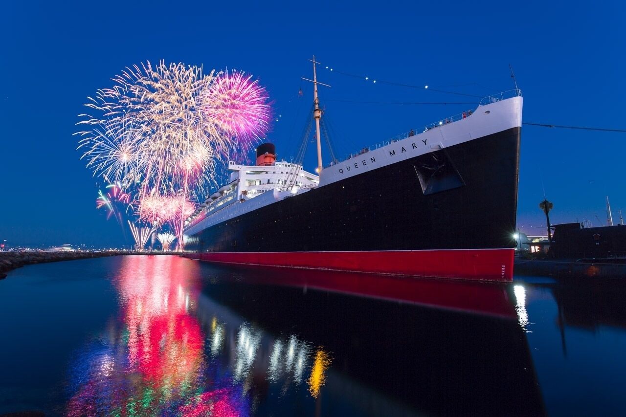 A large cruise ship named "Queen Mary" docked at a port with fireworks in the background.