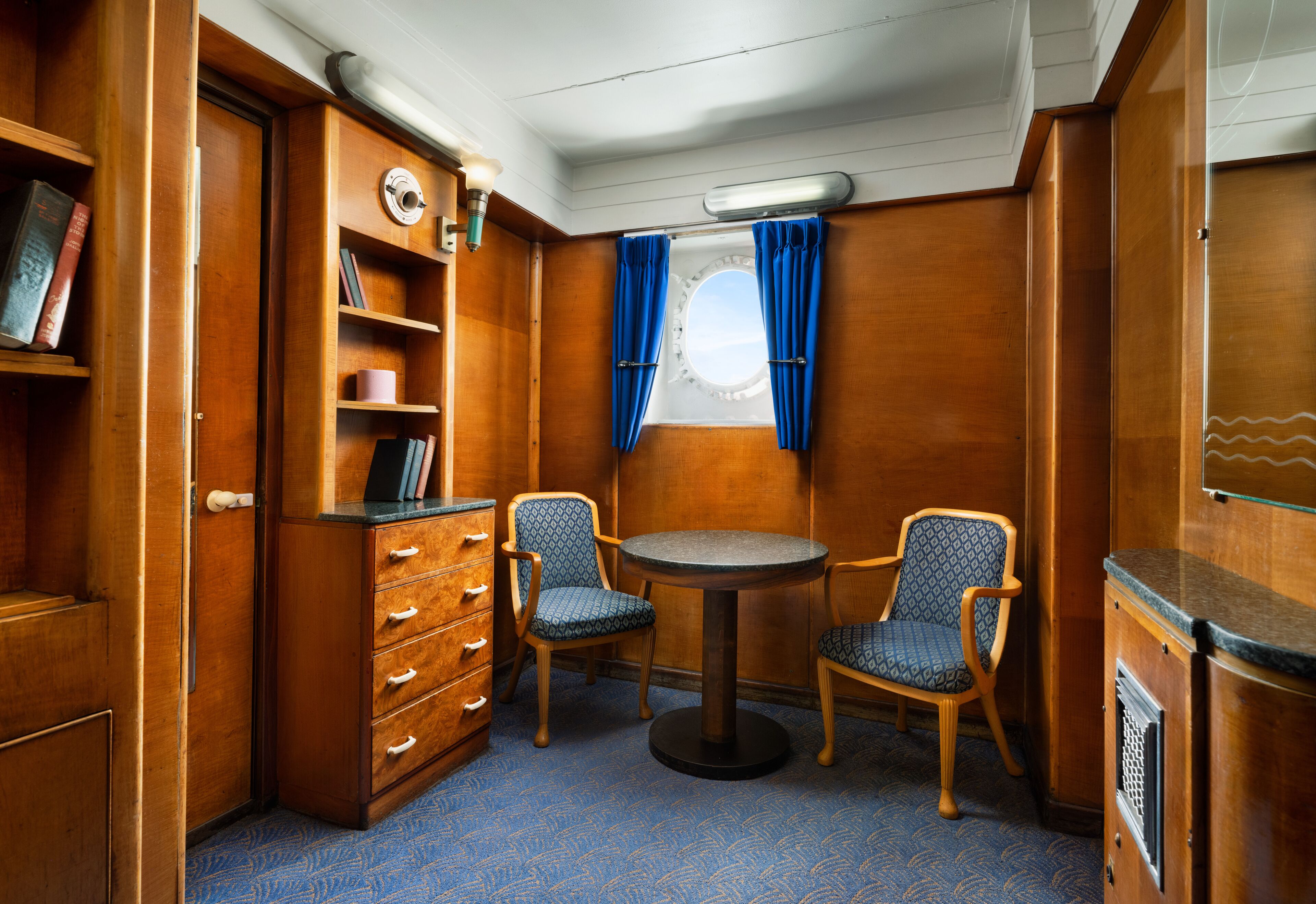 A compact room with a wooden cabinet, a small table with two chairs, a window with curtains, and a bookshelf.