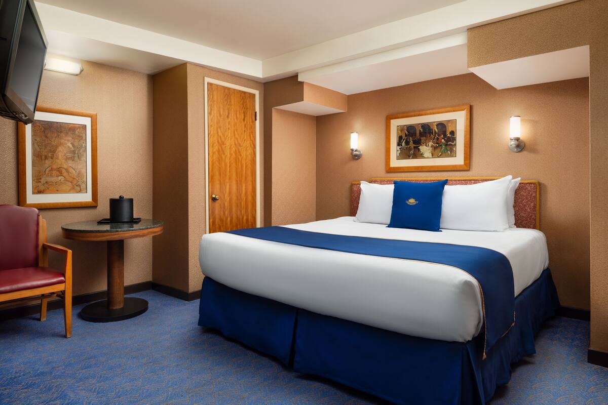 Habitación de hotel con una cama grande, una mesita redonda pequeña, una silla, televisión y obras de arte en las paredes.
