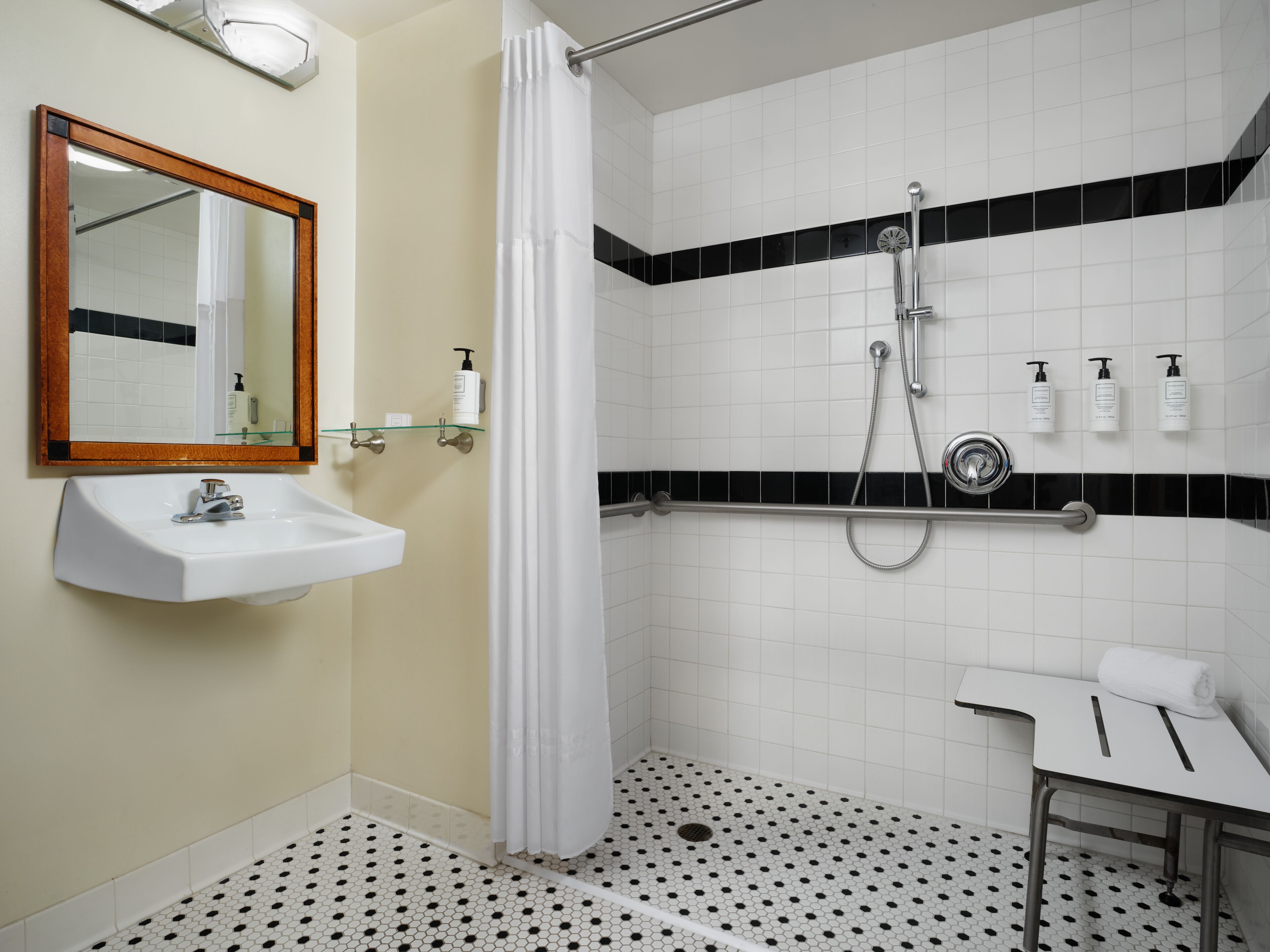 Un baño con lavamanos blanco, espejo y ducha. Hay artículos de aseo en la pared y una toalla en el suelo.