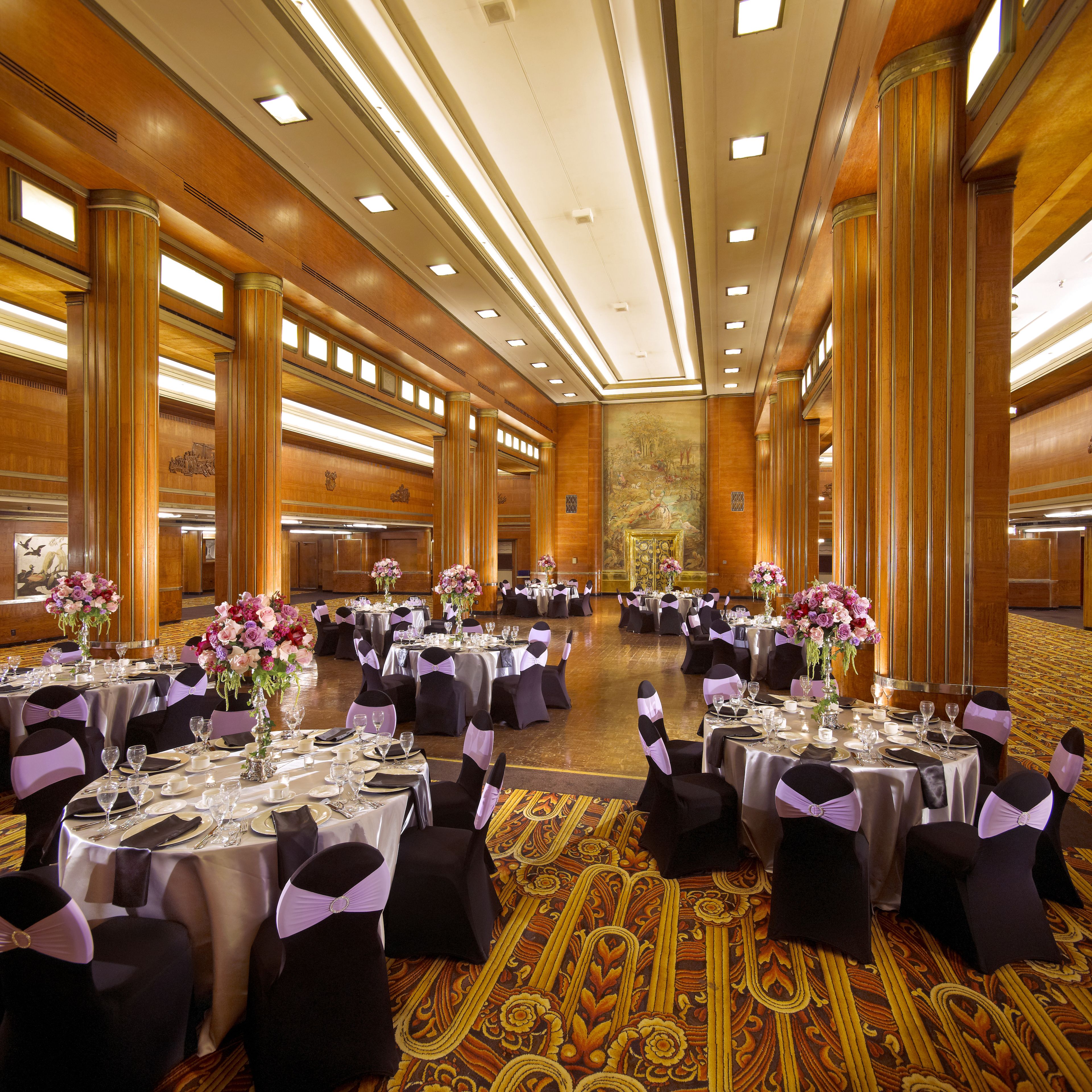 Un amplio salón de banquetes con varias mesas redondas, cada una con mantel blanco, sillas negras y cintas moradas.
