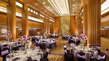 Un amplio salón de banquetes con varias mesas redondas, cada una con mantel blanco, sillas negras y cintas moradas.