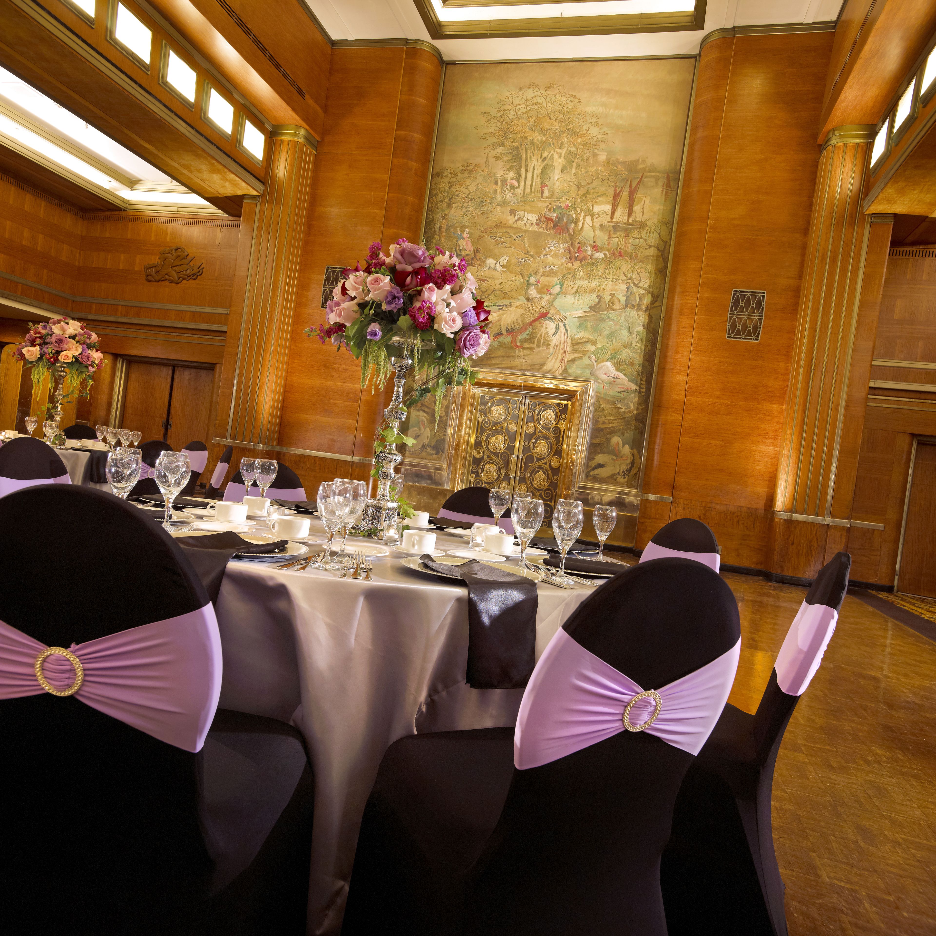 Een feestzaal met een lange tafel gedekt voor een formeel evenement, met aan weerszijden stoelen met paarse linten, een grote bloemstuk in het midden en een muurschildering op de muur.