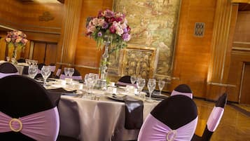 Une salle de banquet avec une longue table dressée pour un événement formel, bordée de chaises ornées de rubans violets, un grand arrangement floral au centre et une fresque murale.