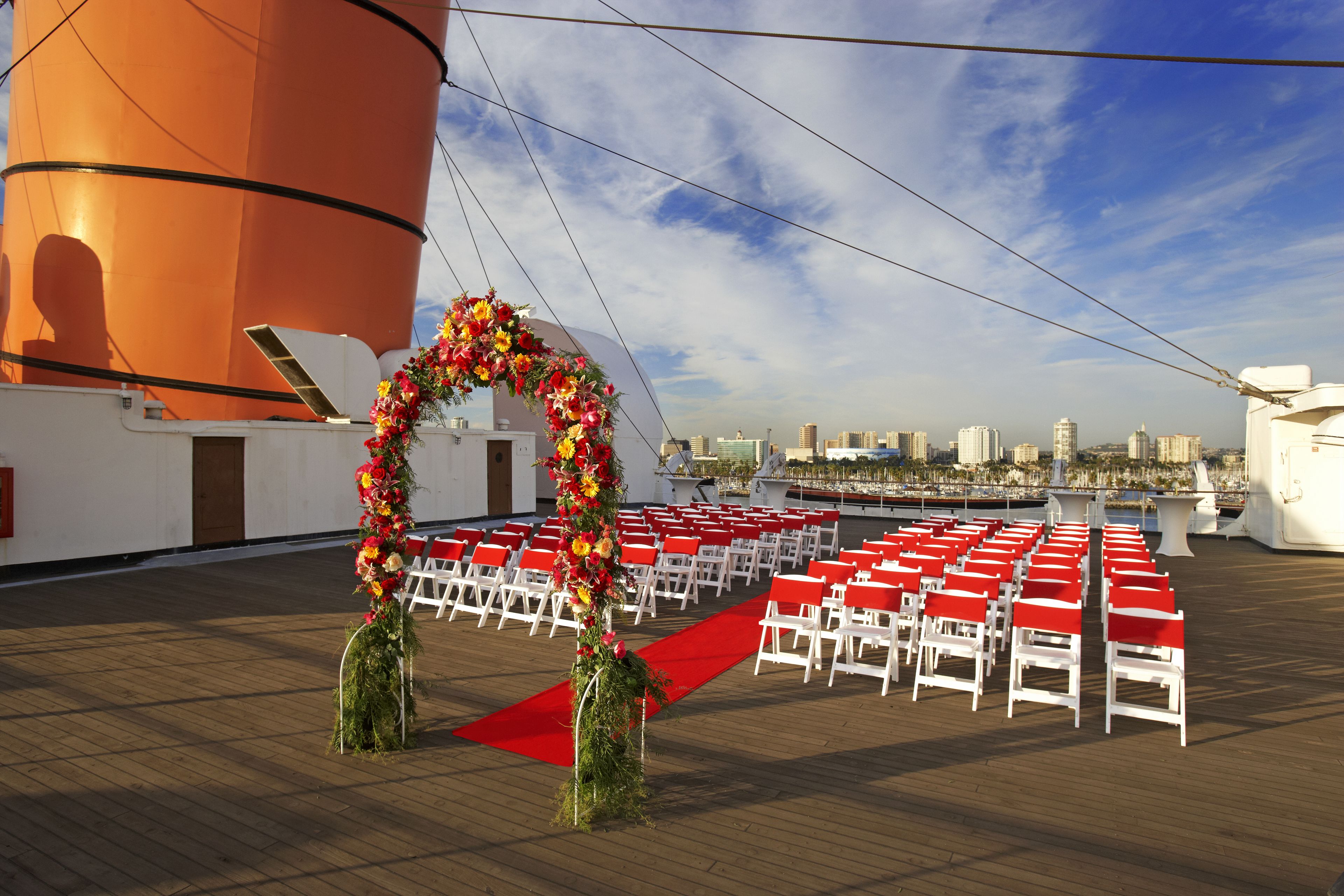 A wedding ceremony setup on a ship deck with a red carpet aisle, white chairs, and a floral arch.
