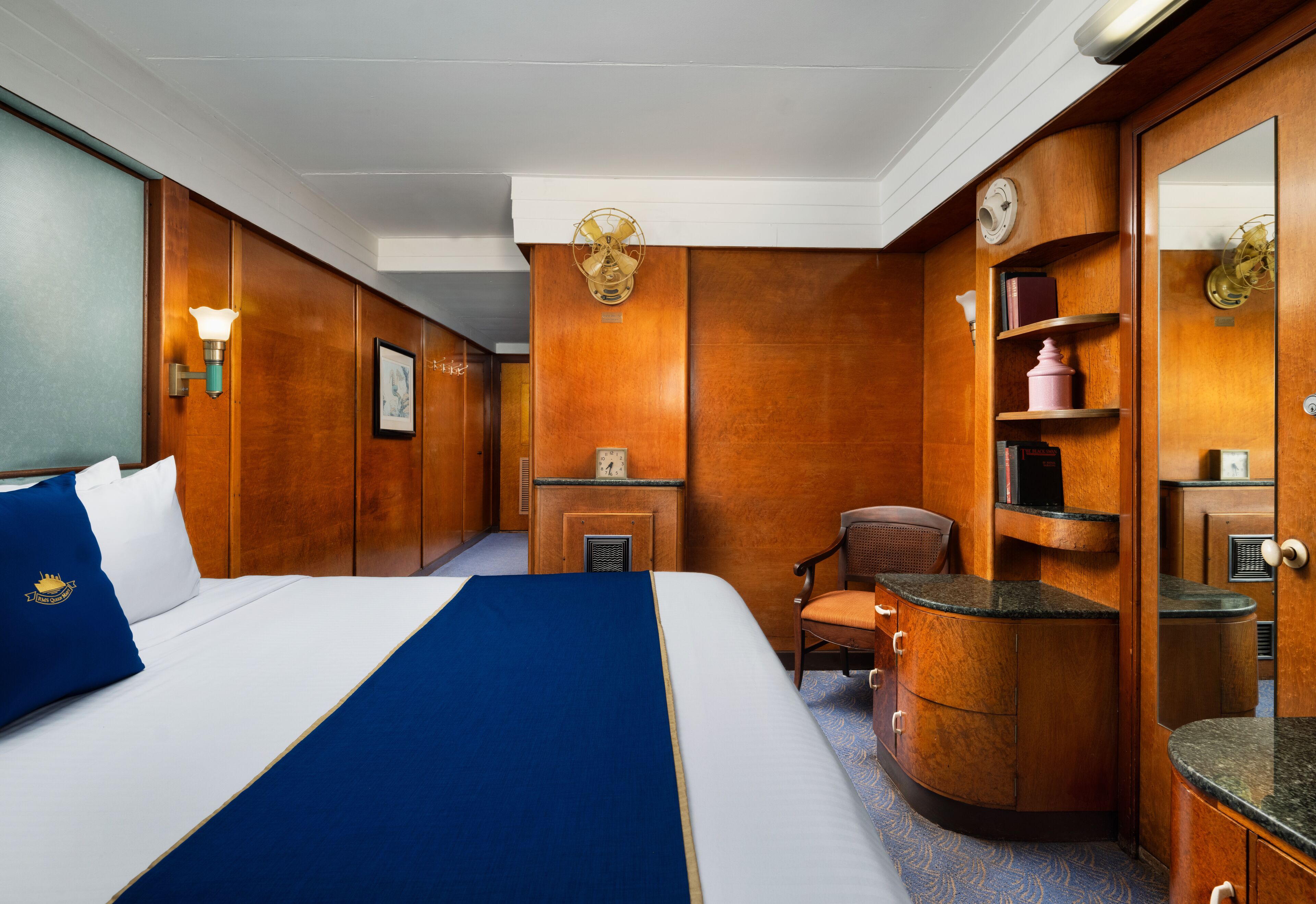 Une chambre d’hôtel avec un grand lit, des boiseries, un bureau, une chaise, un miroir et une étagère avec des livres et un vase.