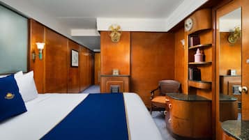Une chambre d’hôtel avec un grand lit, des boiseries, un bureau, une chaise, un miroir et une étagère avec des livres et un vase.