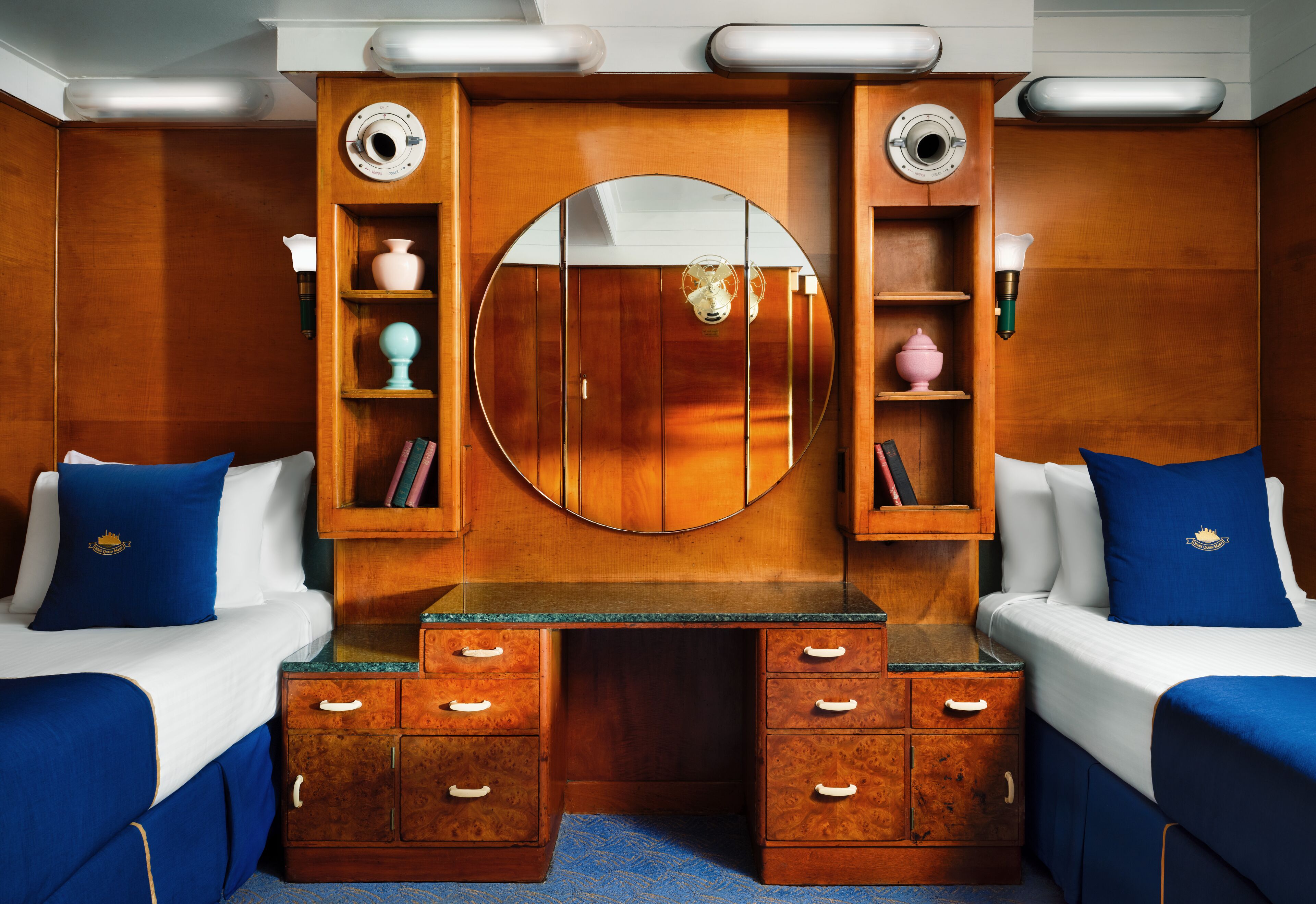 A room with two beds, a wooden vanity with drawers, a large mirror, and shelves with decorative items.