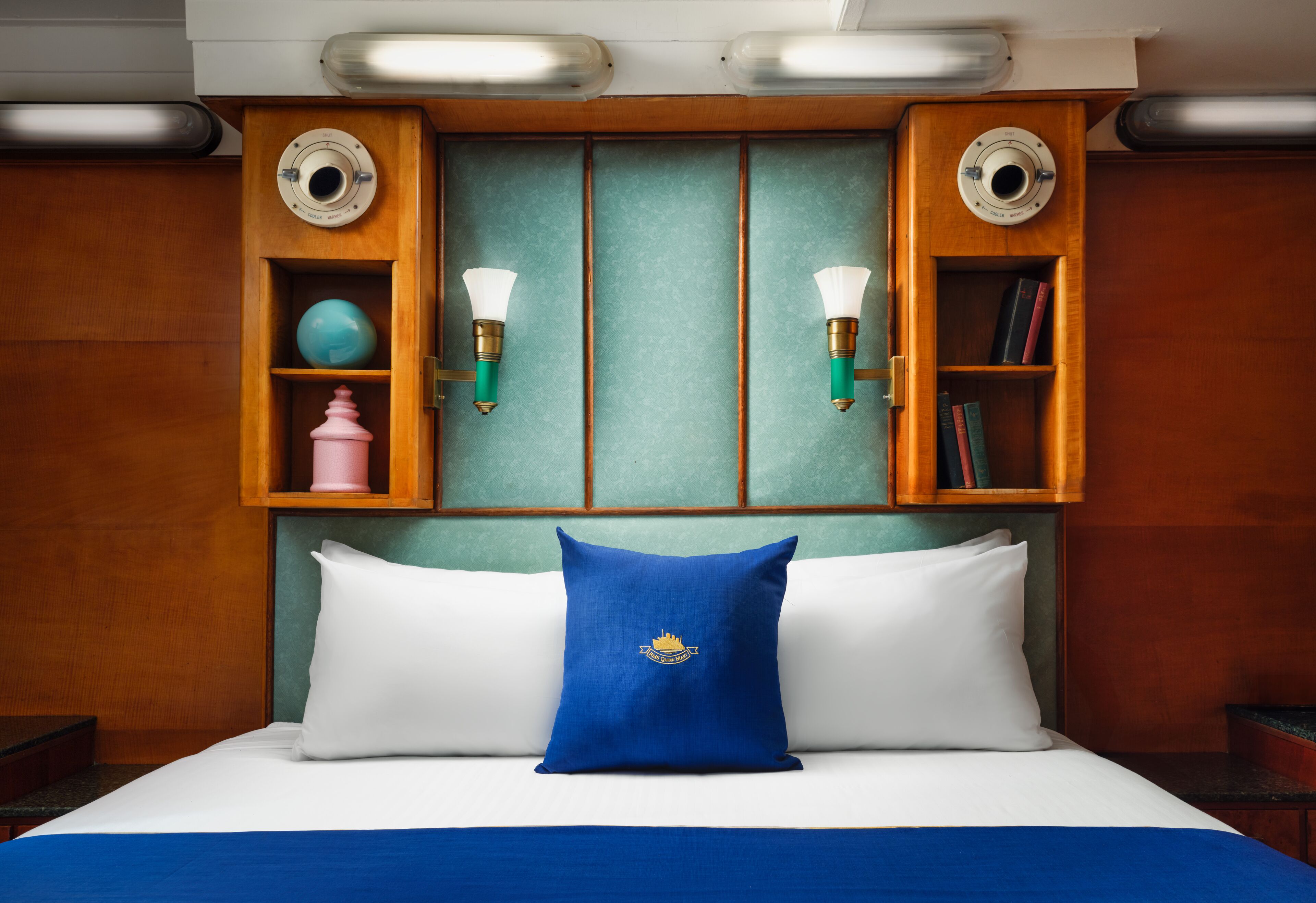 A neatly made bed with a blue pillow, wooden headboard, and two wall-mounted shelves with books and decorative items.