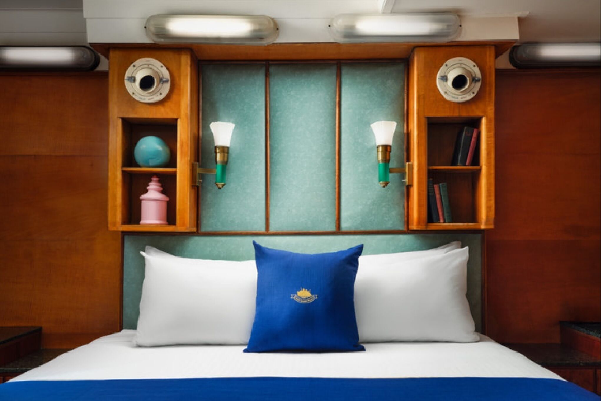 A hotel room with a bed, two wall sconces, a blue pillow, and a shelf with decorative items.