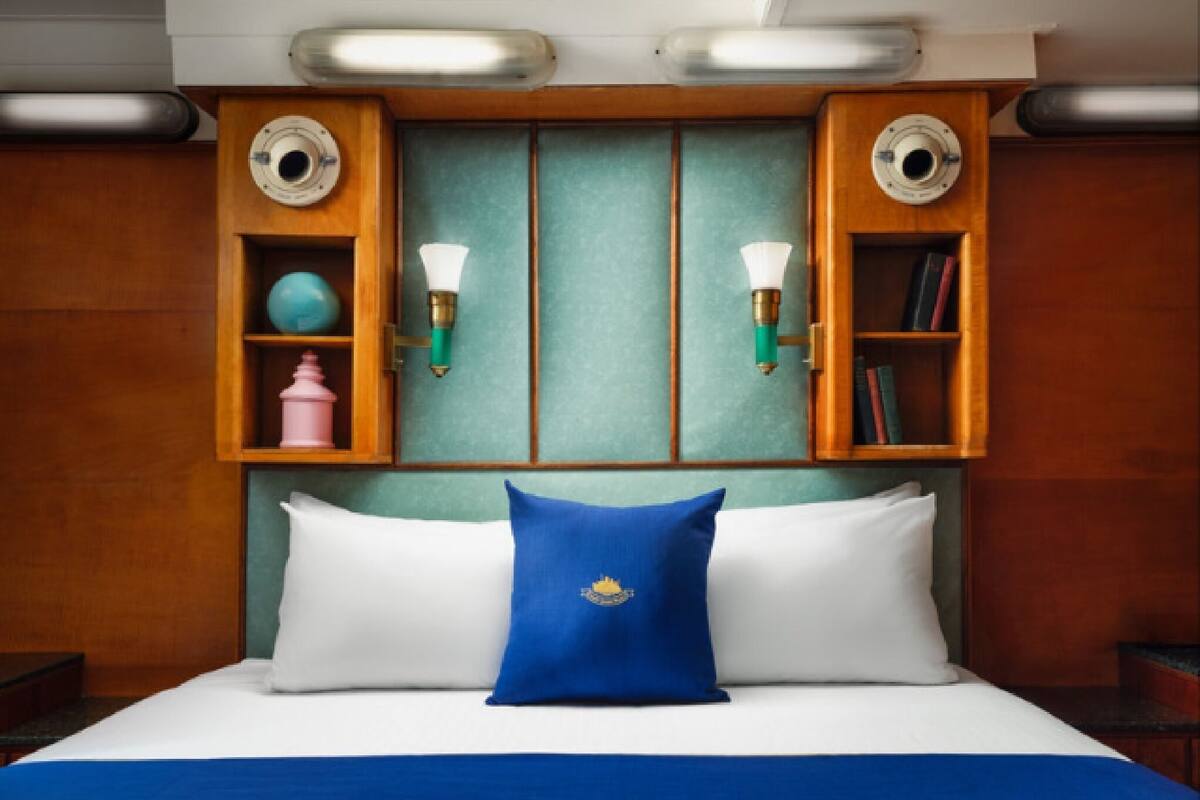 A hotel room with a bed, two wall sconces, a blue pillow, and a shelf with decorative items.