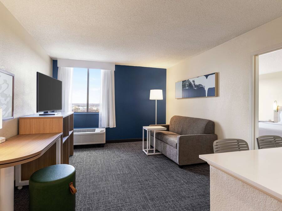 Quarto de hotel com escrivaninha, televisão, sofá, mesa pequena e janela com cortinas.