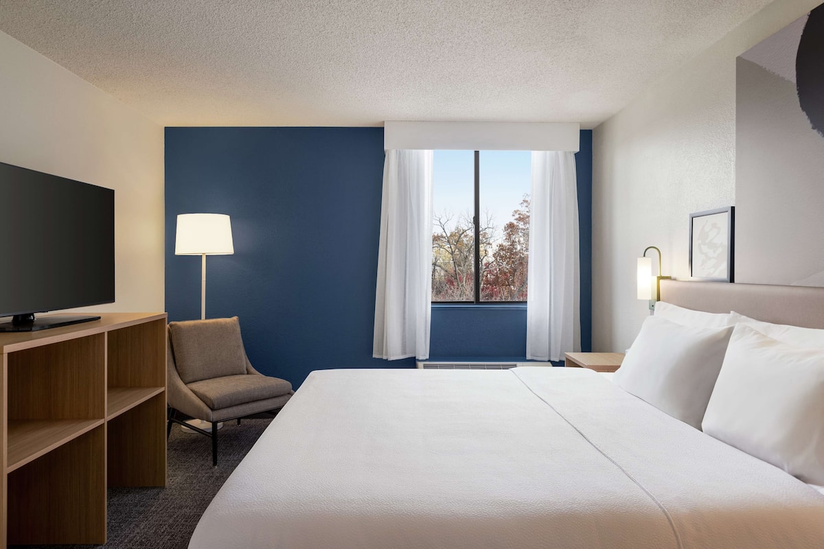 Ein Hotelzimmer mit einem großen Bett, einem Fernseher auf einem Holzboden, einem Sessel und einem Fenster mit Vorhängen.