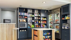 A modern hotel kitchen with built-in appliances, shelves stocked with snacks and beverages, and a wooden counter. - Spark by Hilton Germantown Washington DC North (Germantown)