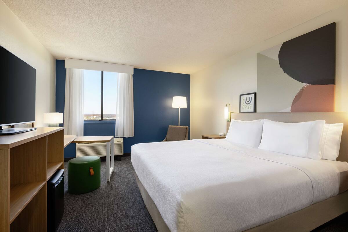 Ein Hotelzimmer mit Bett, Schreibtisch, Stuhl, Lampe und einem Fenster mit Vorhängen.
