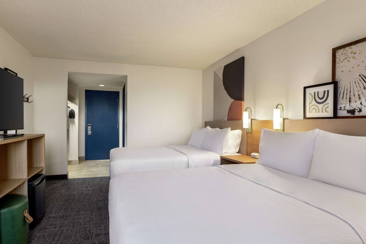 Ein Hotelzimmer mit zwei Betten, einem Fernseher, einem Schreibtisch und einer Tür zu einem Flur.