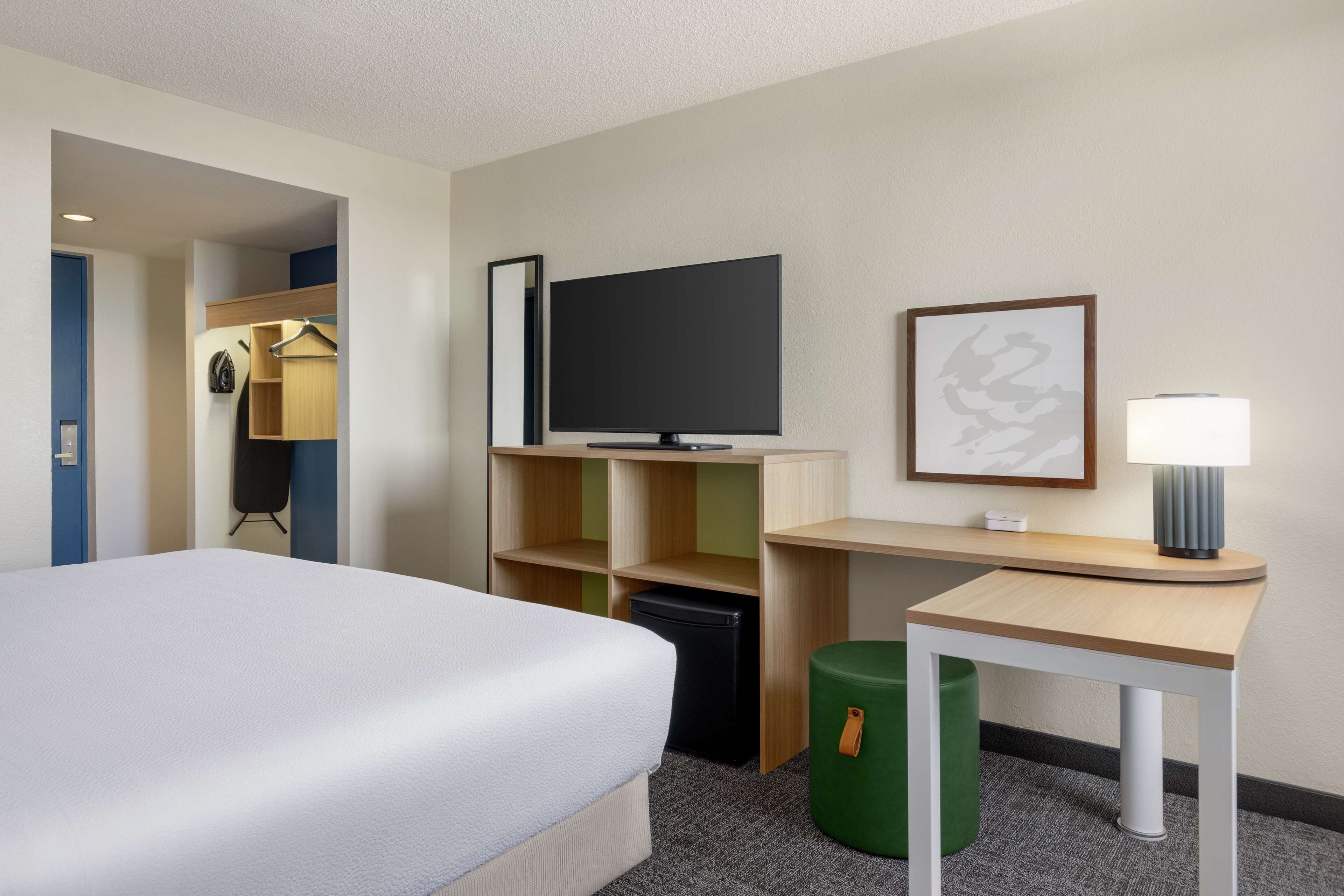 A hotel room with a bed, a desk, a television, a lamp, a trash bin, and a wardrobe.