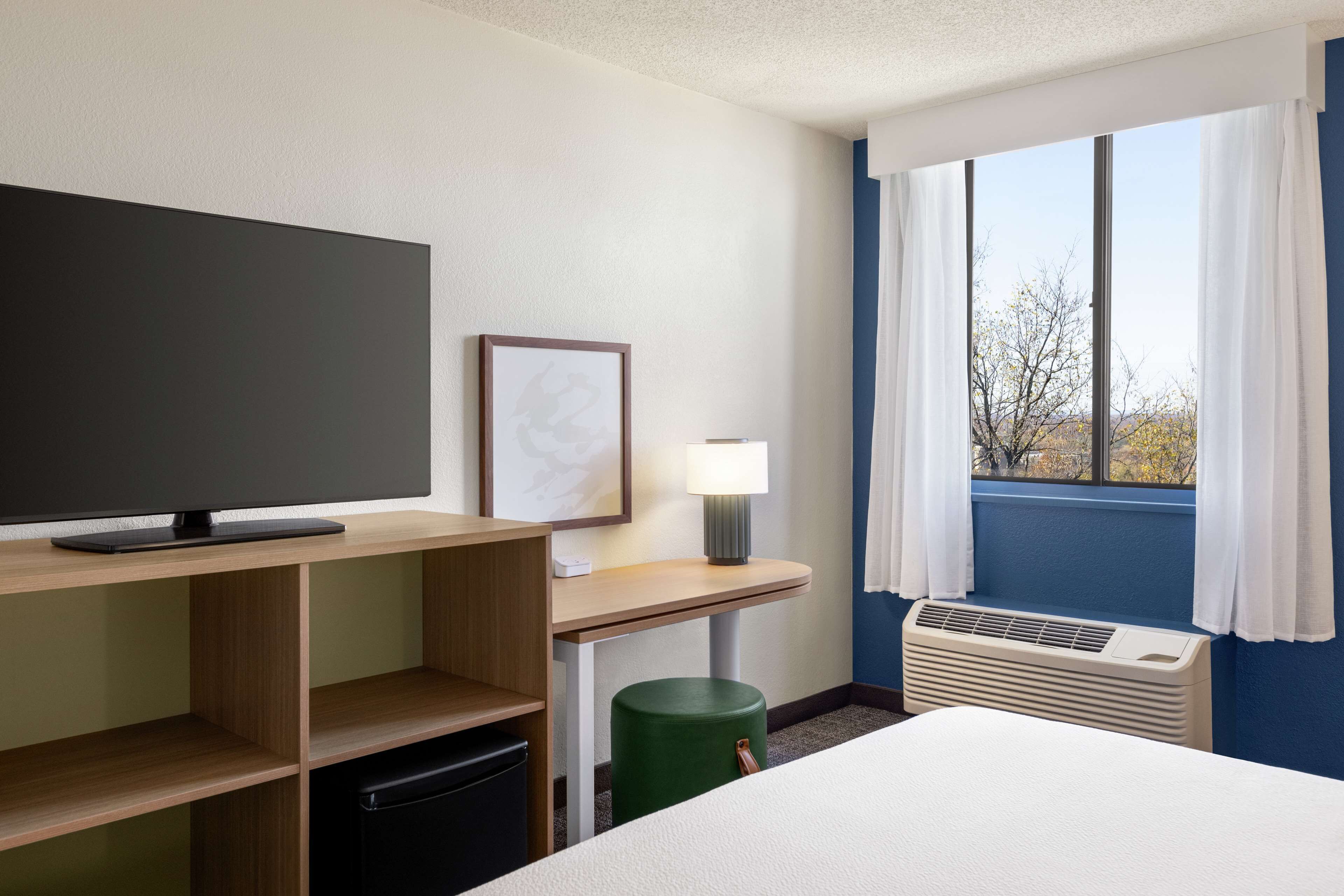A hotel room with a bed, a desk with a flat-screen TV, a lamp, a heater, and a window with curtains.
