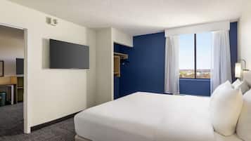 A hotel room with a large bed, a flat-screen TV, a window with curtains, and a view of the city.