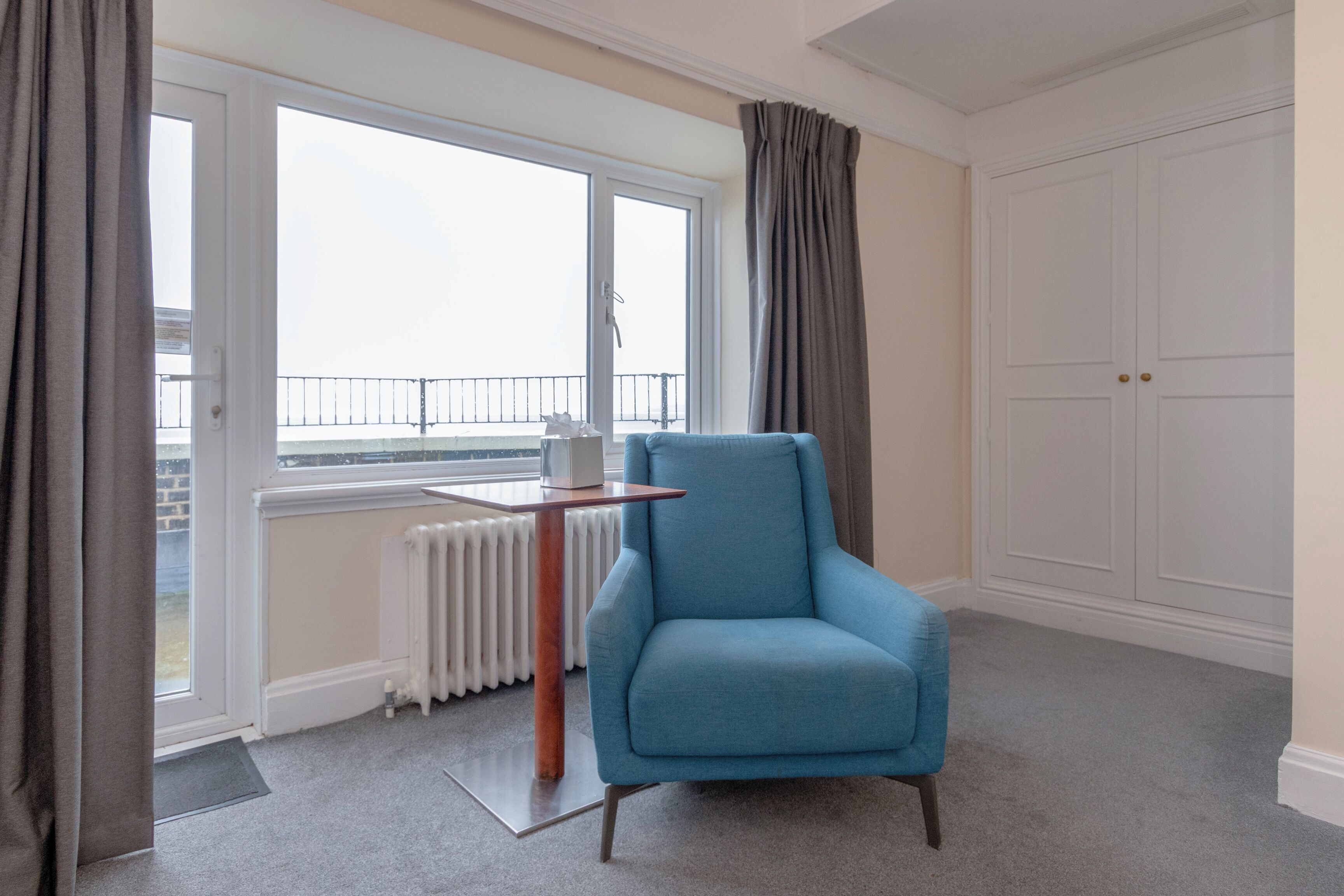 Ein Zimmer mit blauem Sessel, Heizkörper, einem fenster mit Vorhängen und einem weißen Kleiderschrank.