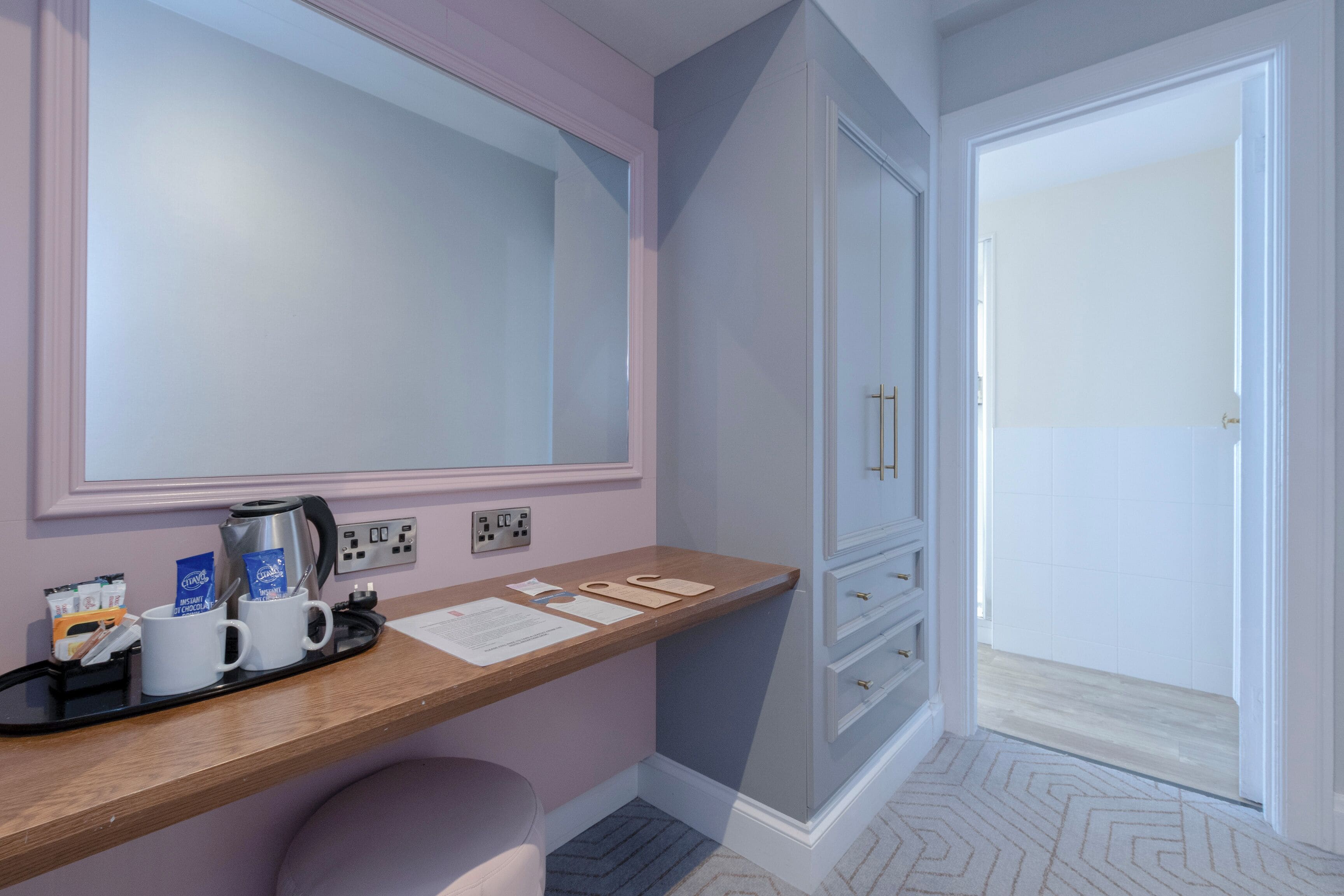 A hotel room with a wooden desk, a mirror, a wardrobe, and a door leading to a bathroom.