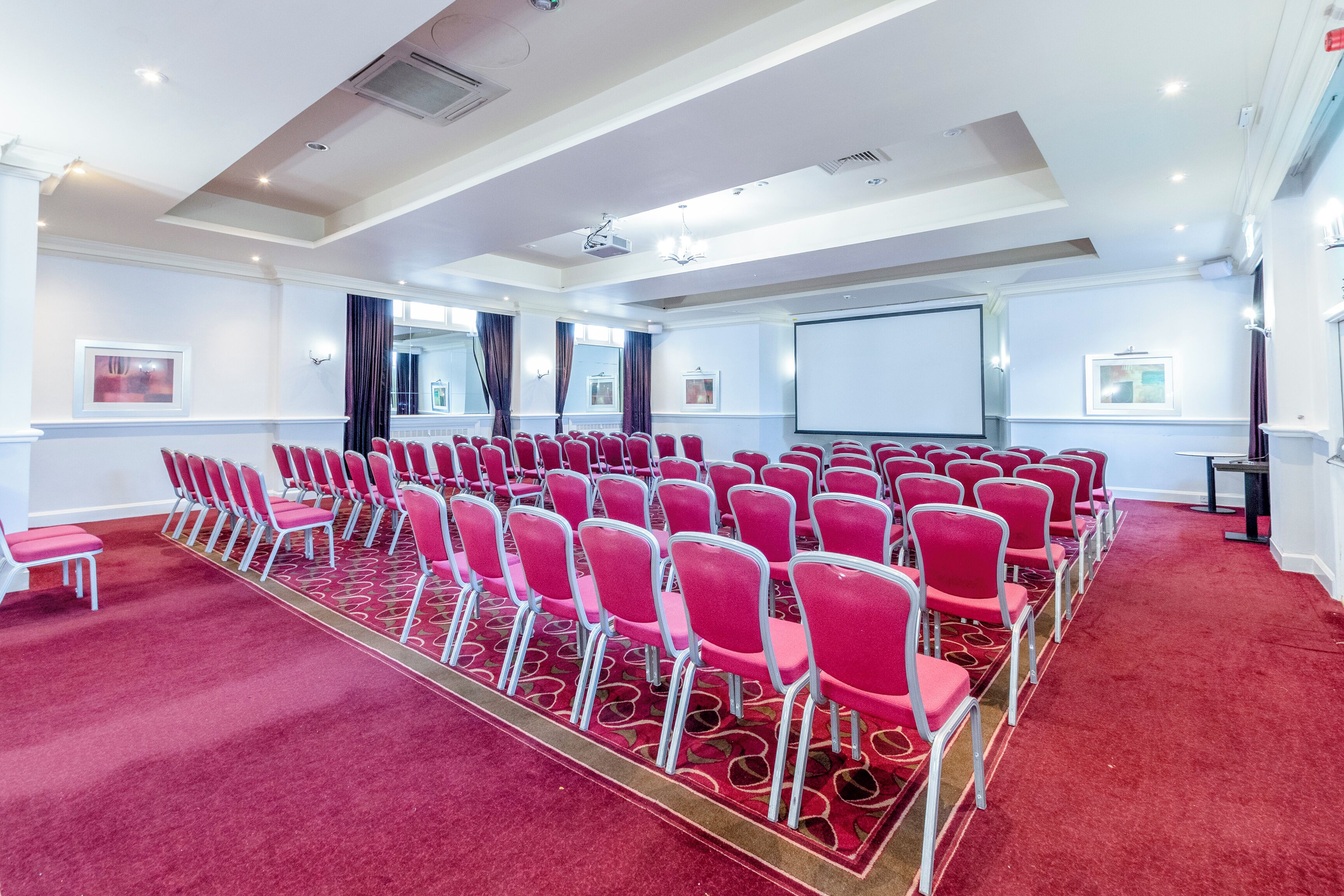 A conference room with rows of red chairs, a projector screen, and a carpeted floor.