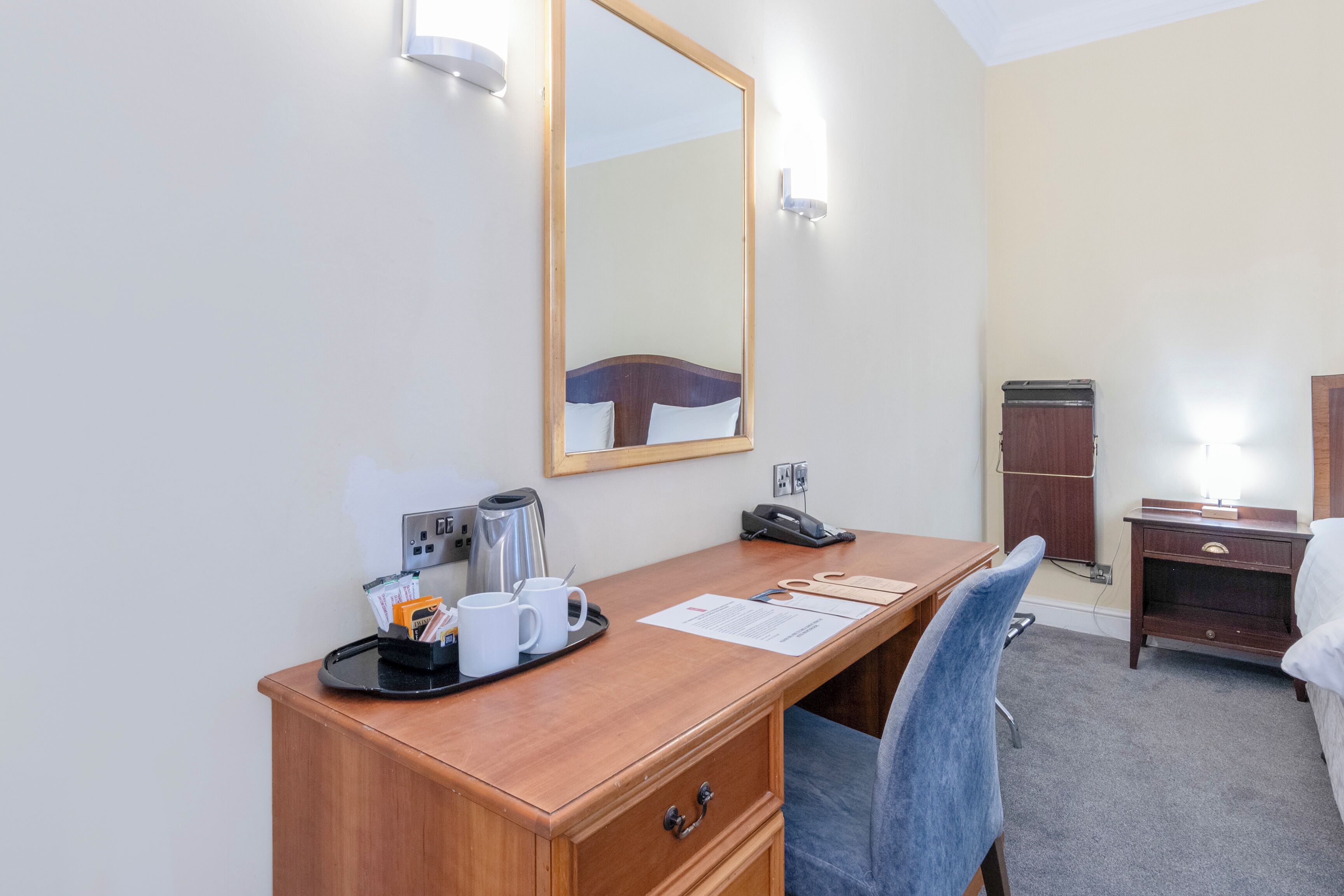 Habitación de hotel con un escritorio de madera, una silla, una cama, una mesita de noche, un espejo, un enchufe, un teléfono, una bandeja con tazas y una cafetera, y un armario pequeño.