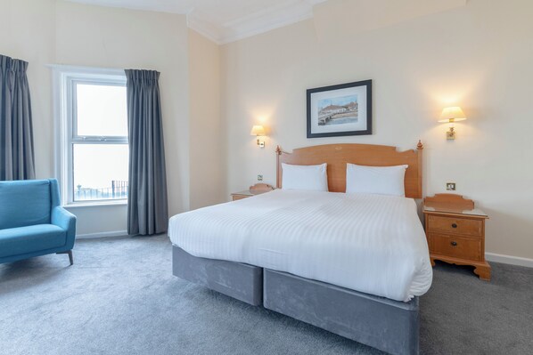 A hotel room with a large bed, a blue armchair, a window with curtains, two bedside tables with lamps, and a framed picture on the wall.