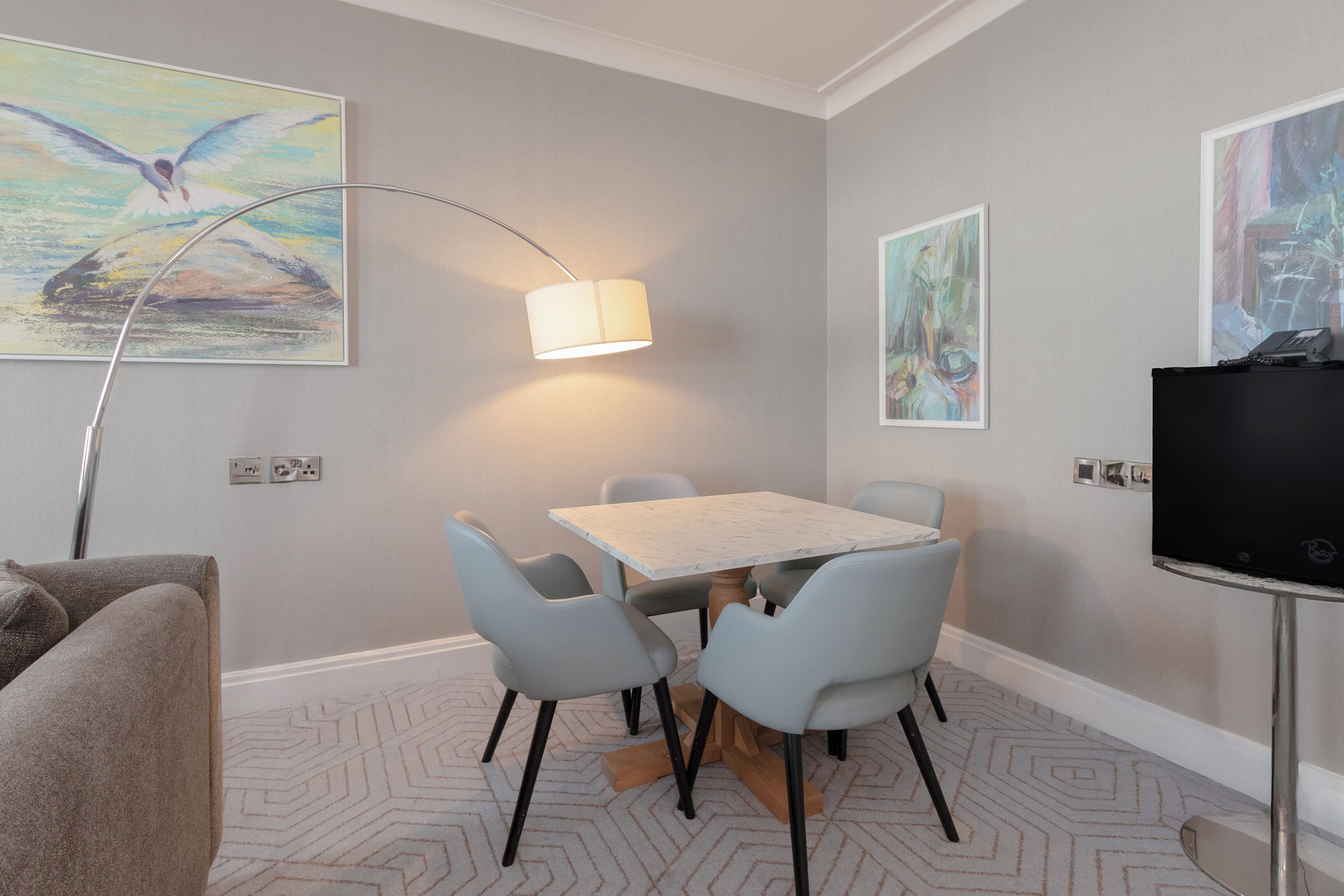 A room with a dining table, four chairs, a lamp, a television, and artwork on the walls.