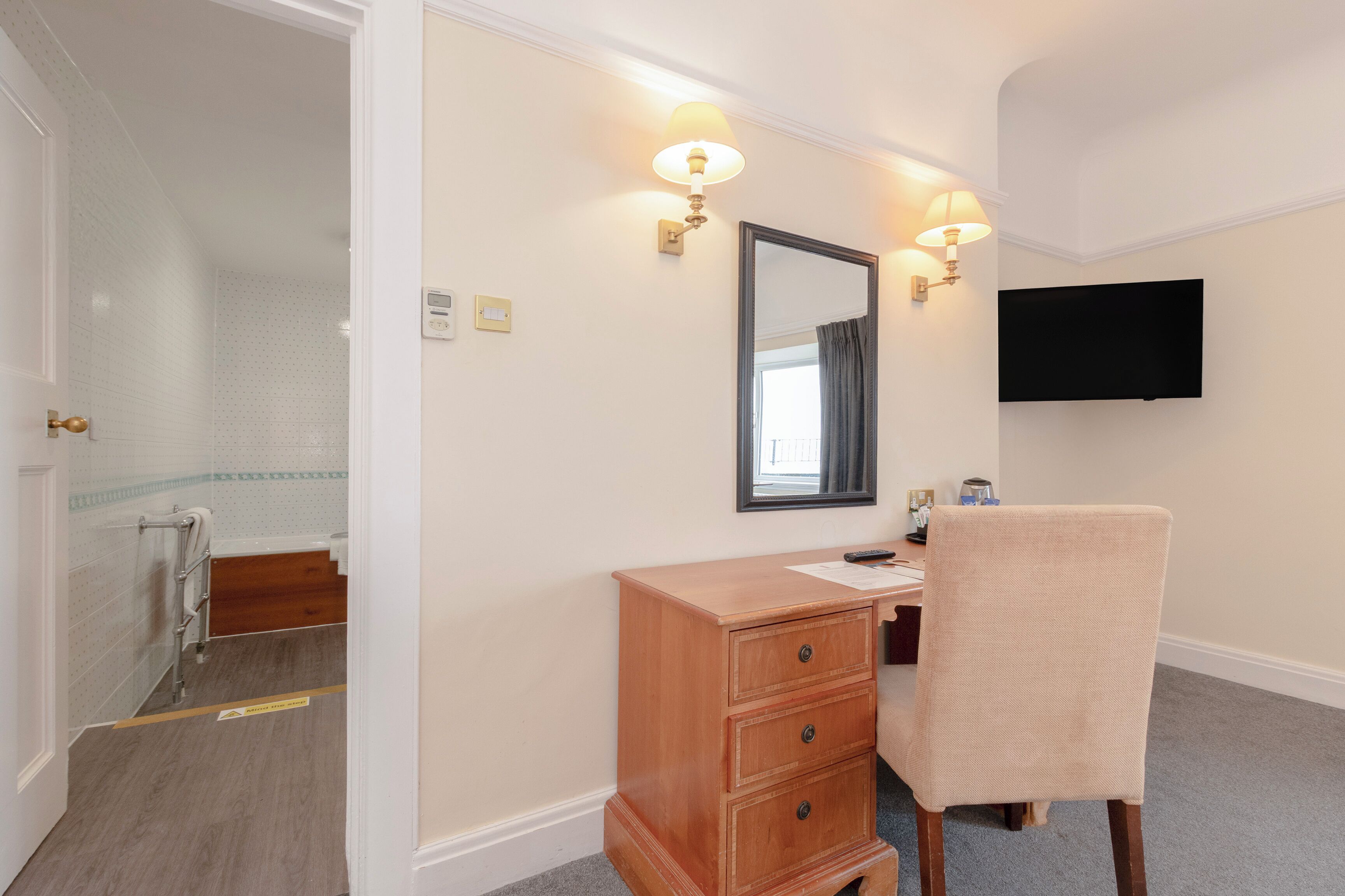 Una habitación de hotel con un aparador de madera, una silla, un televisor montado en la pared y un baño visible a través de una puerta abierta.