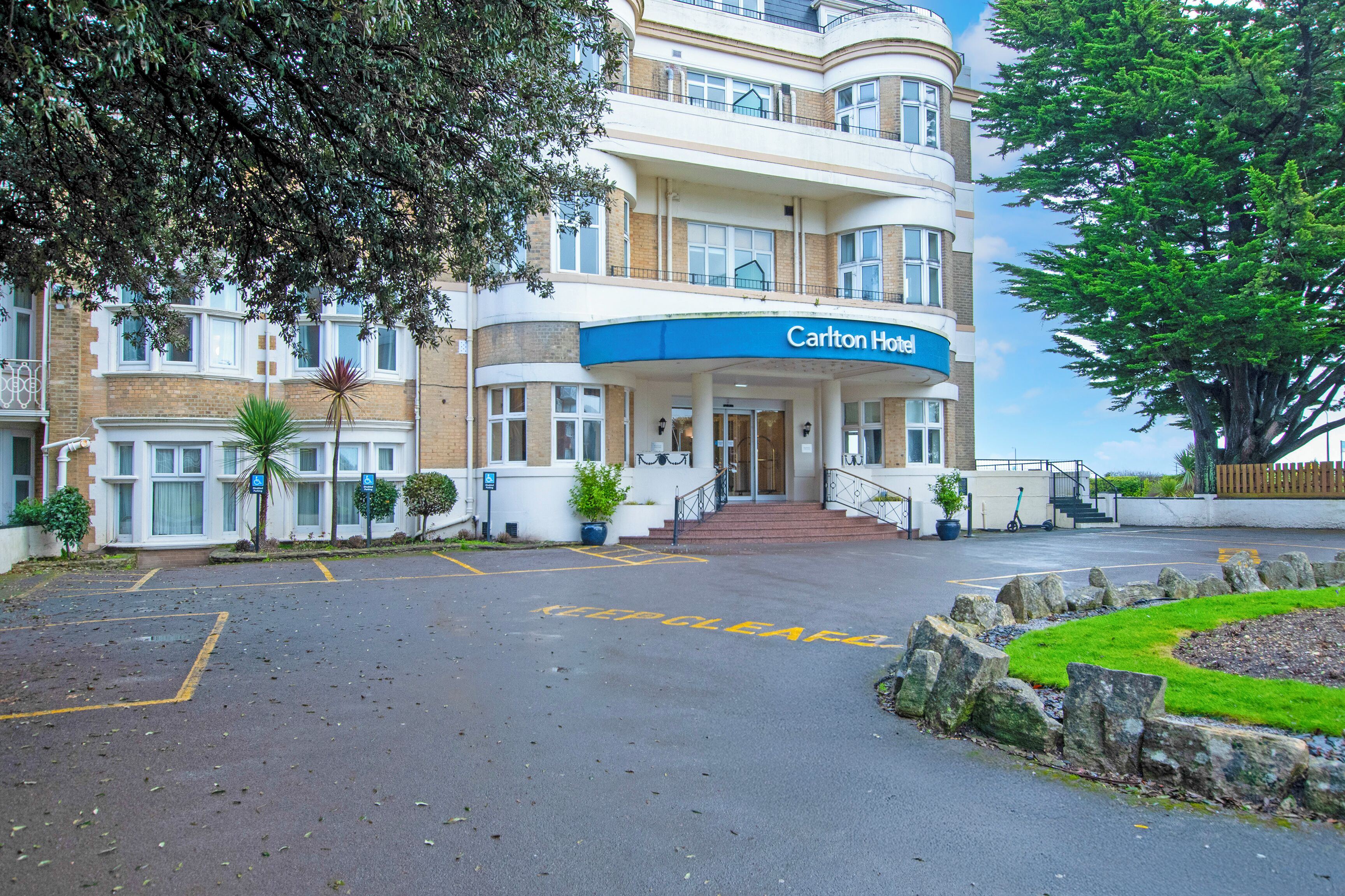 A hotel entrance with a sign reading 'Carlton Hotel,' a parking area, and a landscaped garden.