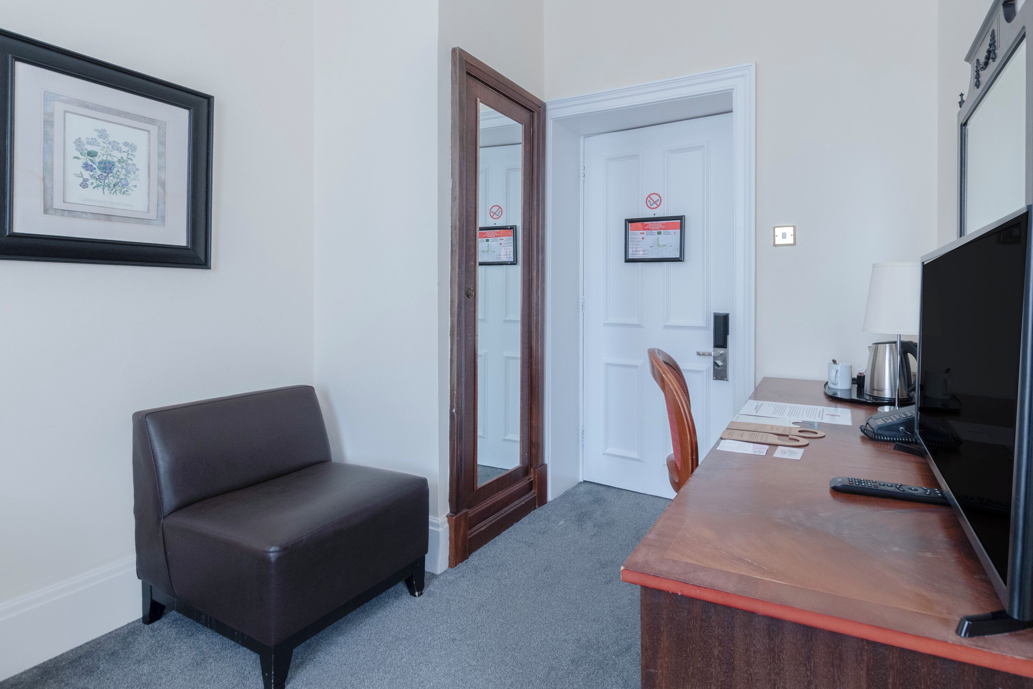 A room with a dark brown sofa, a wooden desk with a computer monitor, a remote control, and a framed picture on the wall.
