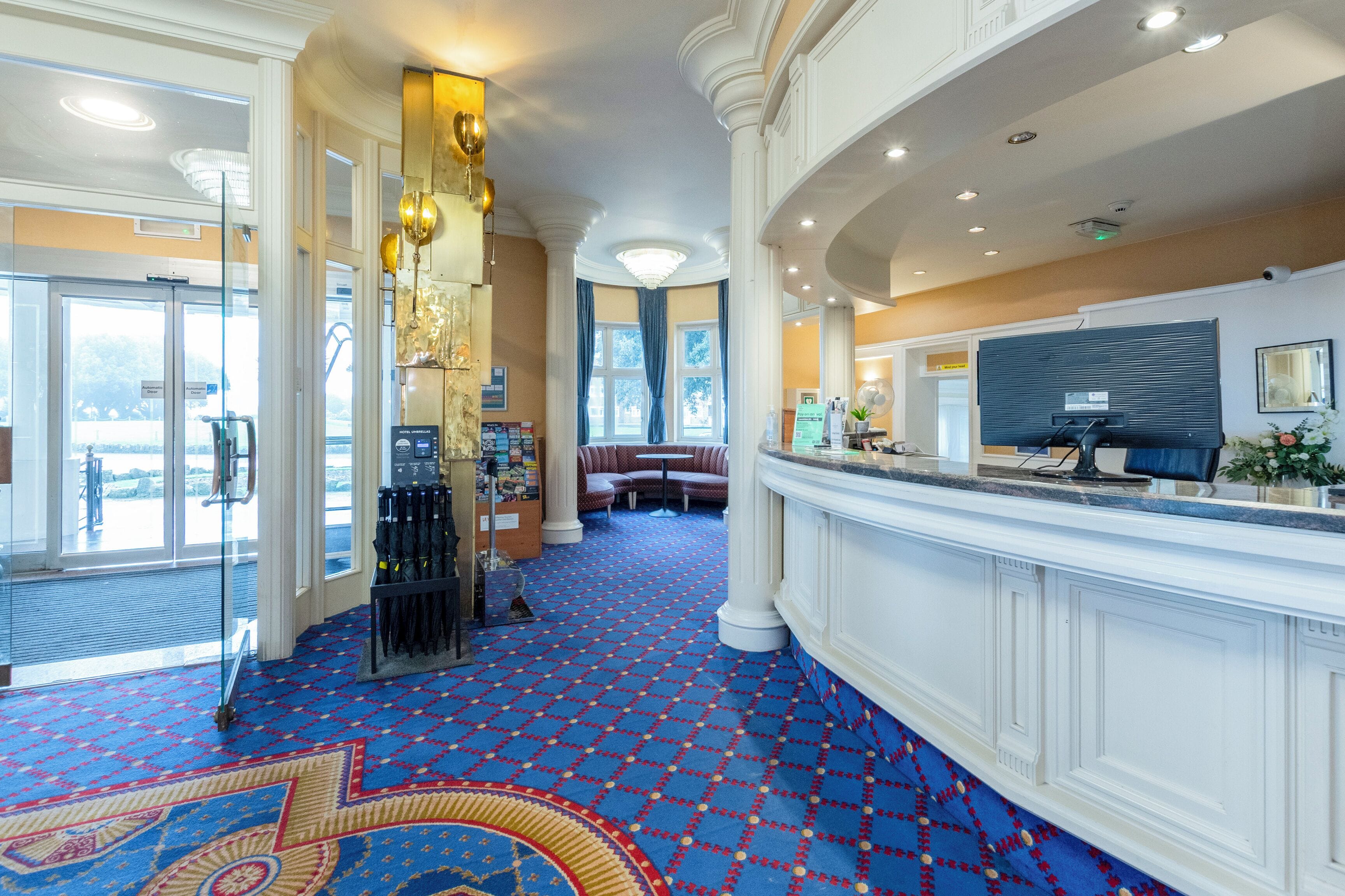A hotel lobby with a patterned carpet, a reception desk, a seating area, and a large window.