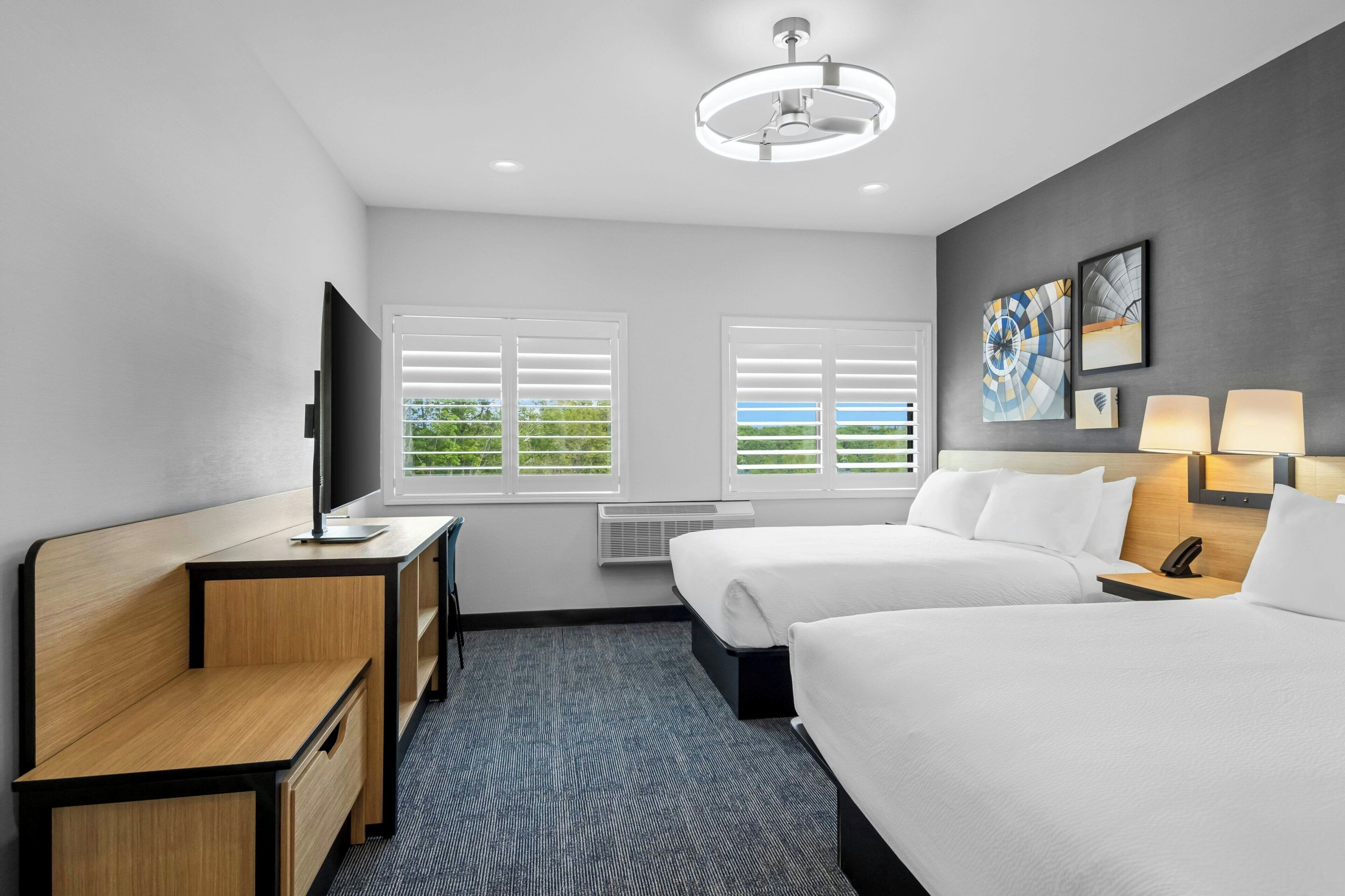 A hotel room with a desk, two beds, a TV, a ceiling fan, and artwork on the wall.