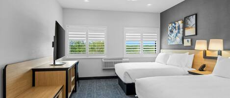 A hotel room with a desk, two beds, a TV, a ceiling fan, and artwork on the wall.