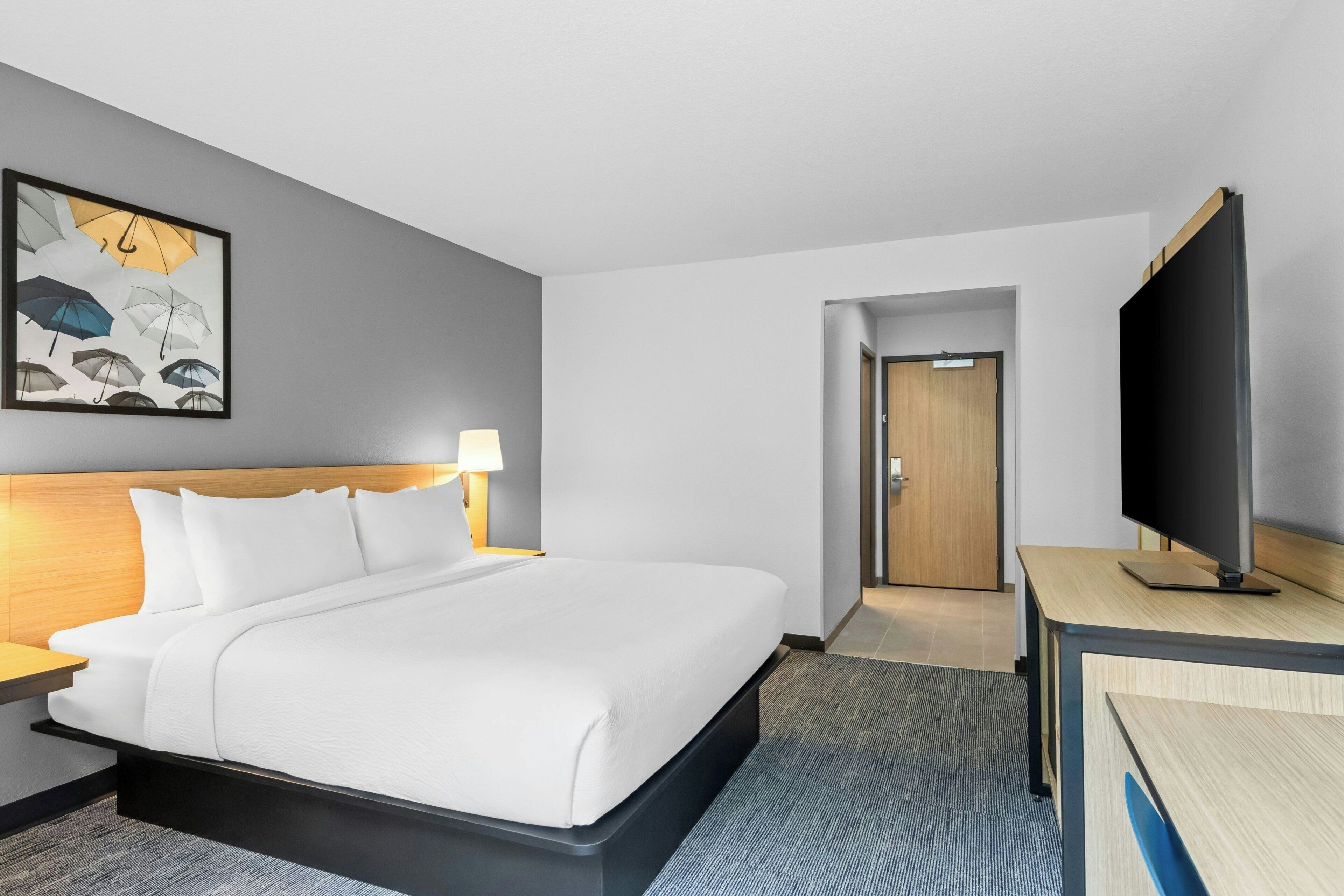 A hotel room with a bed, a desk with a flat-screen TV, a painting of umbrellas, and a door leading to another room.