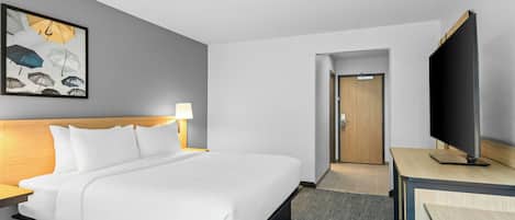 A hotel room with a bed, a desk with a flat-screen TV, a painting of umbrellas, and a door leading to another room.