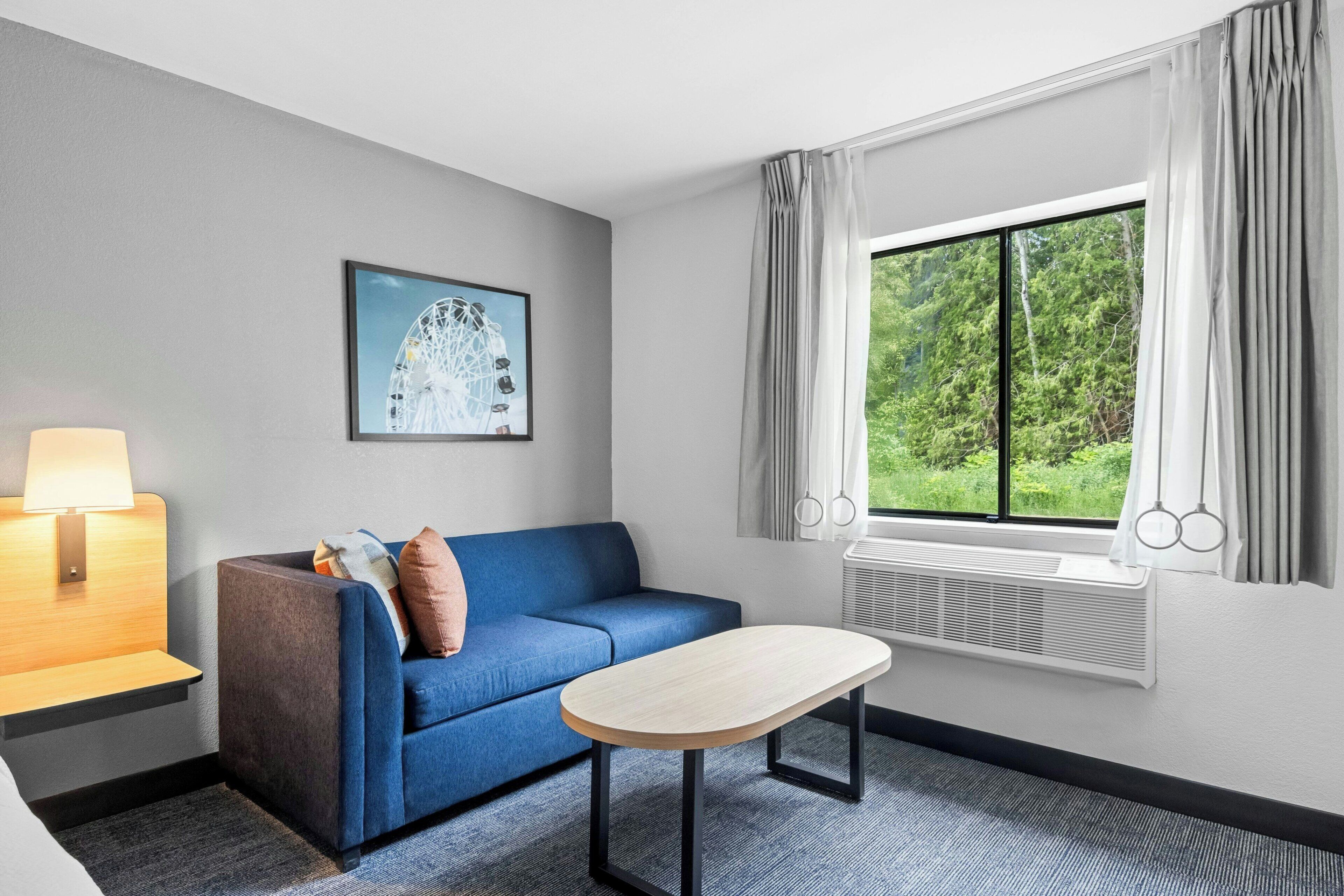 A hotel room with a blue sofa, a small table, a lamp, a picture of a Ferris wheel, and a window with curtains.