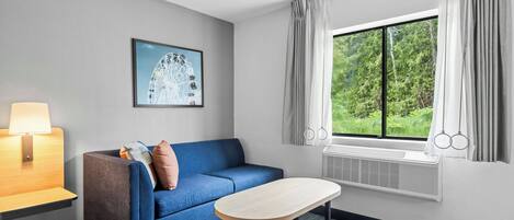 Una habitación de hotel con un sofá azul, una mesita, una lámpara, un cuadro de una noria y una ventana con cortinas.