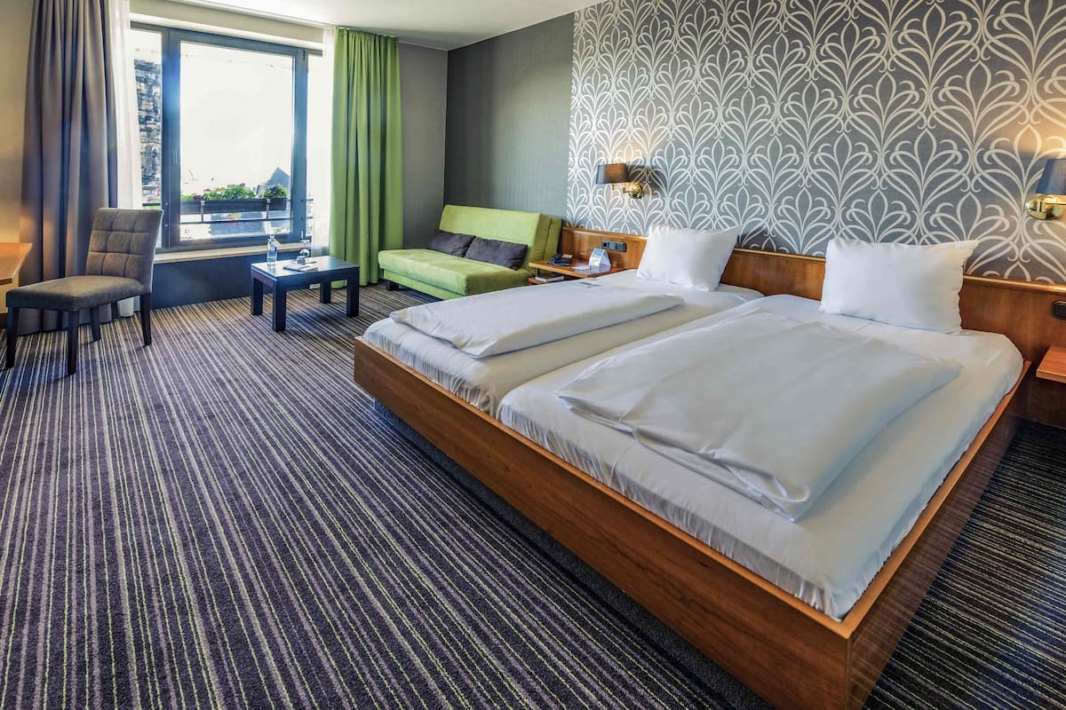 Habitación de hotel con una cama grande, un sofá, una silla, una mesita y una ventana con cortinas.