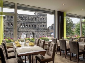 A restaurant with a view of a historic stone building, tables set with yellow napkins, and a modern interior design. - Mercure Hotel Trier Porta Nigra (Trier)