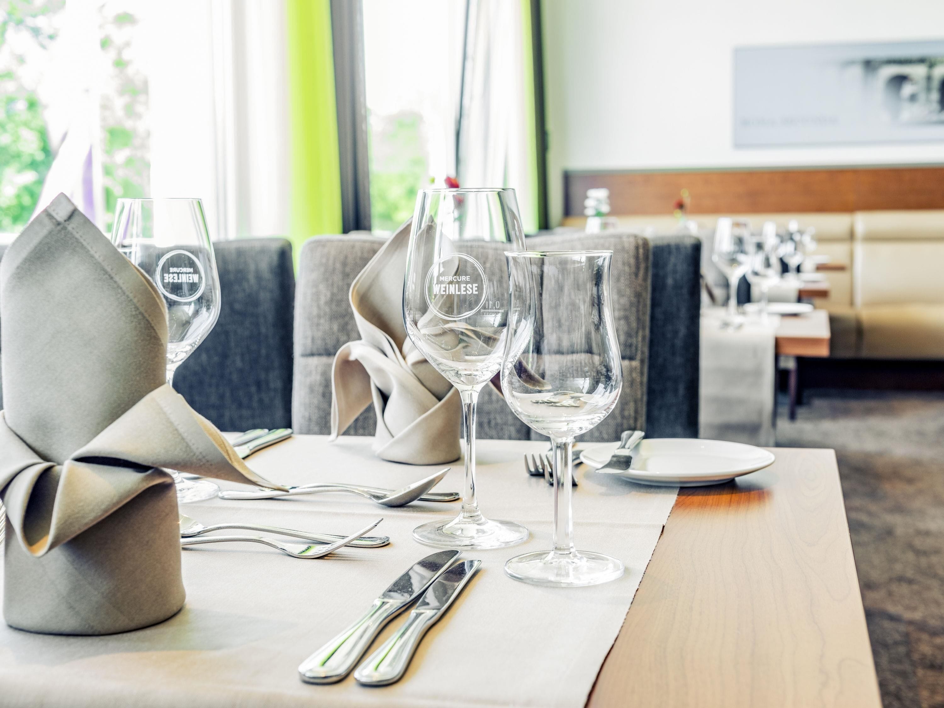 Une table à manger dressée avec des verres à vin, des couverts et un porte-serviettes, dans une chambre d’hôtel moderne donnant sur un paysage verdoyant.