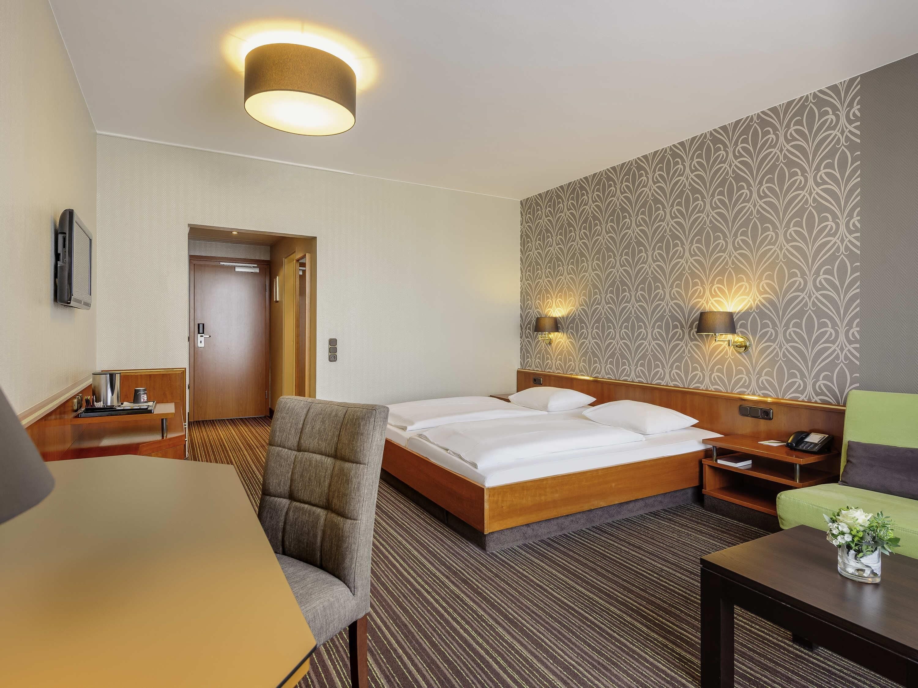A hotel room with a bed, a desk, a chair, a bedside table, a lamp, and a wall with a patterned wallpaper.