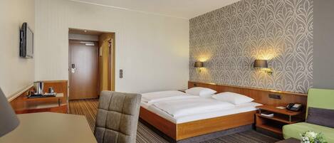 A hotel room with a bed, a desk, a chair, a bedside table, a lamp, and a wall with a patterned wallpaper.