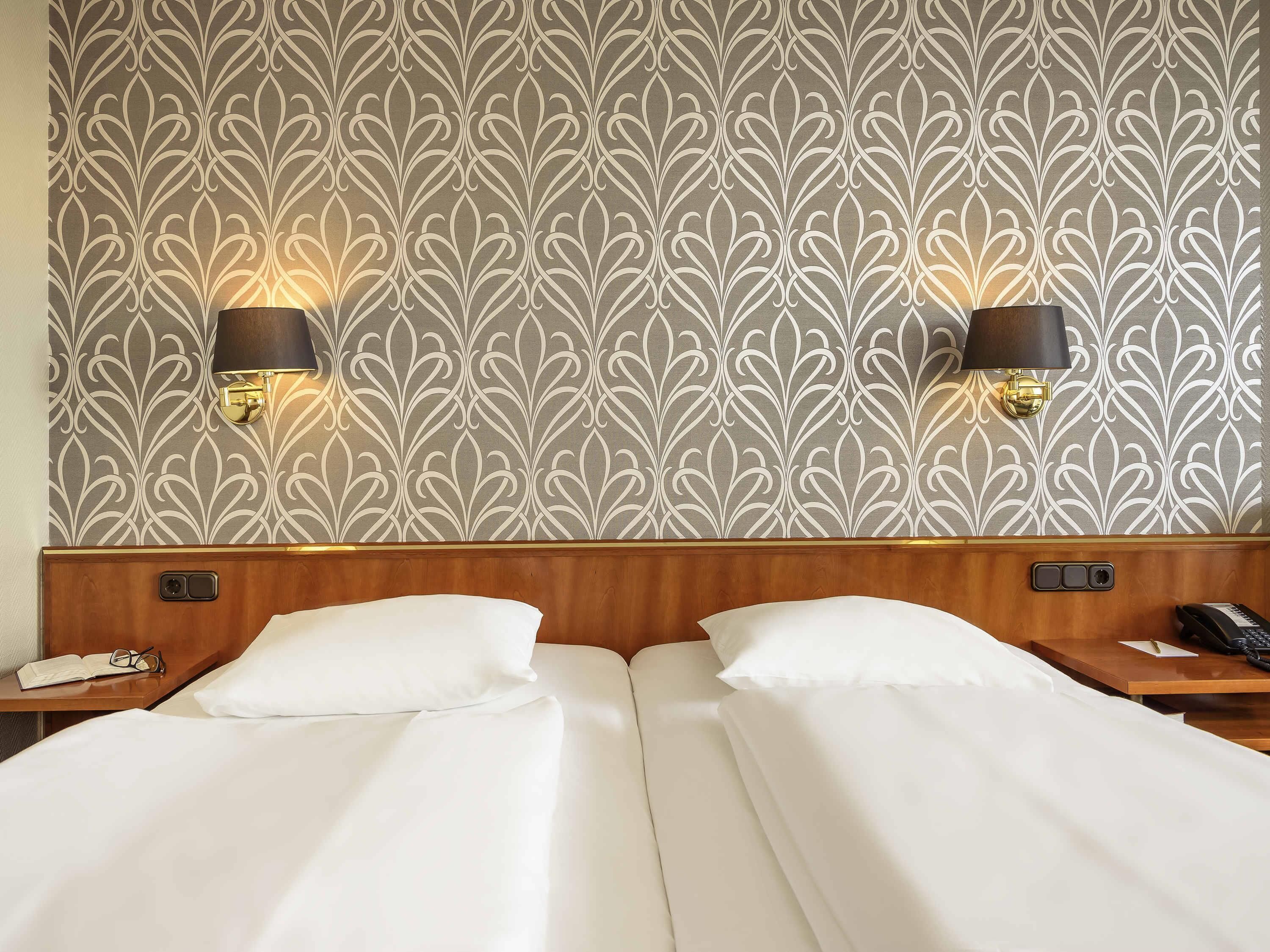 Une chambre d’hôtel avec un papier peint à motifs, deux lampes de chevet, une tête de lit en bois et un lit recouvert de draps blancs.